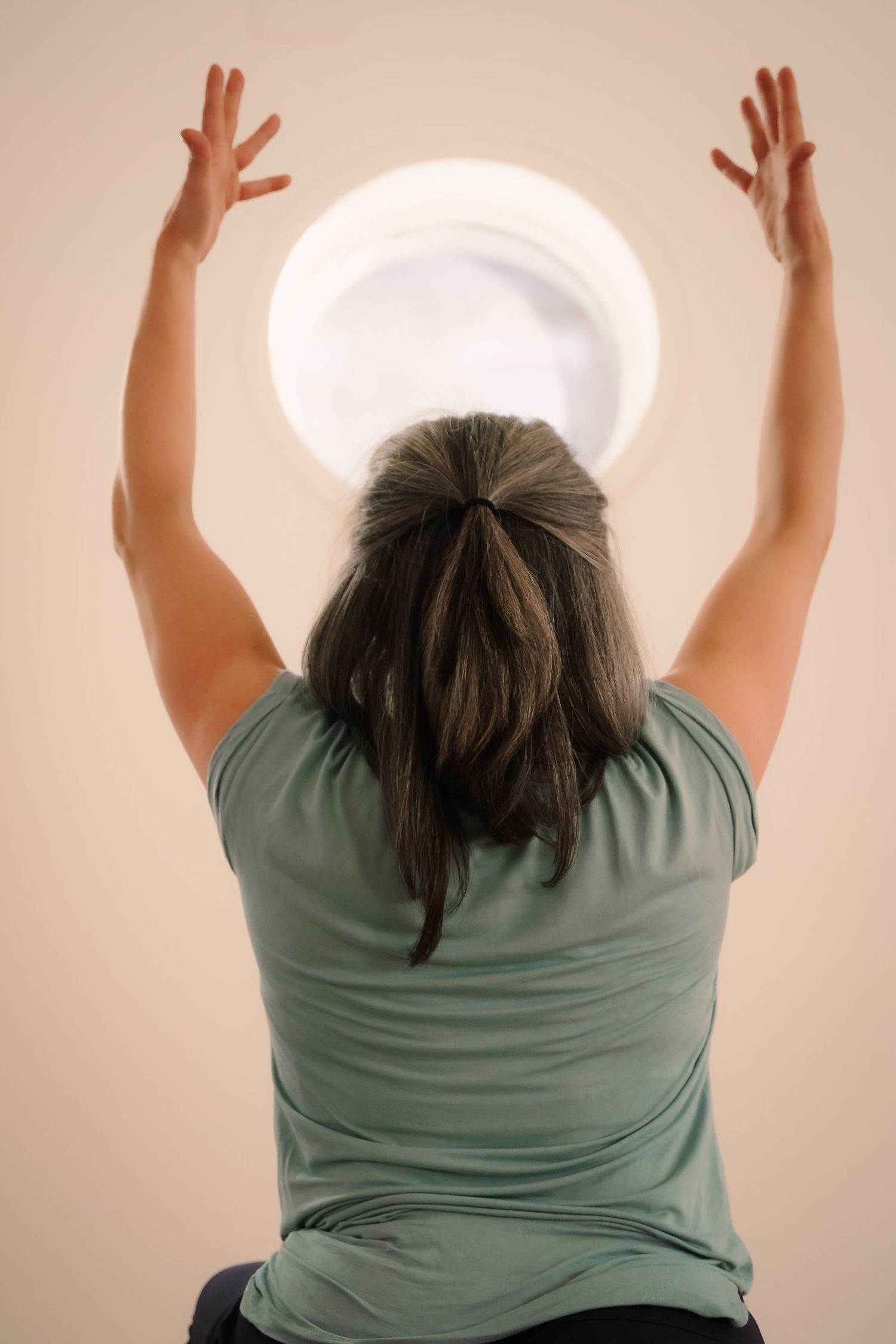 Woman in green top reaching arms overhead toward ceiling light during a yoga or movement class.