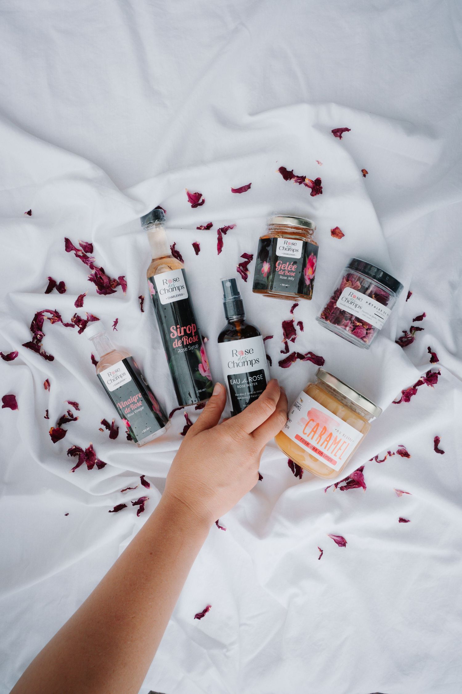 Hand reaching for rose-themed beauty products arranged on white fabric scattered with red rose petals.