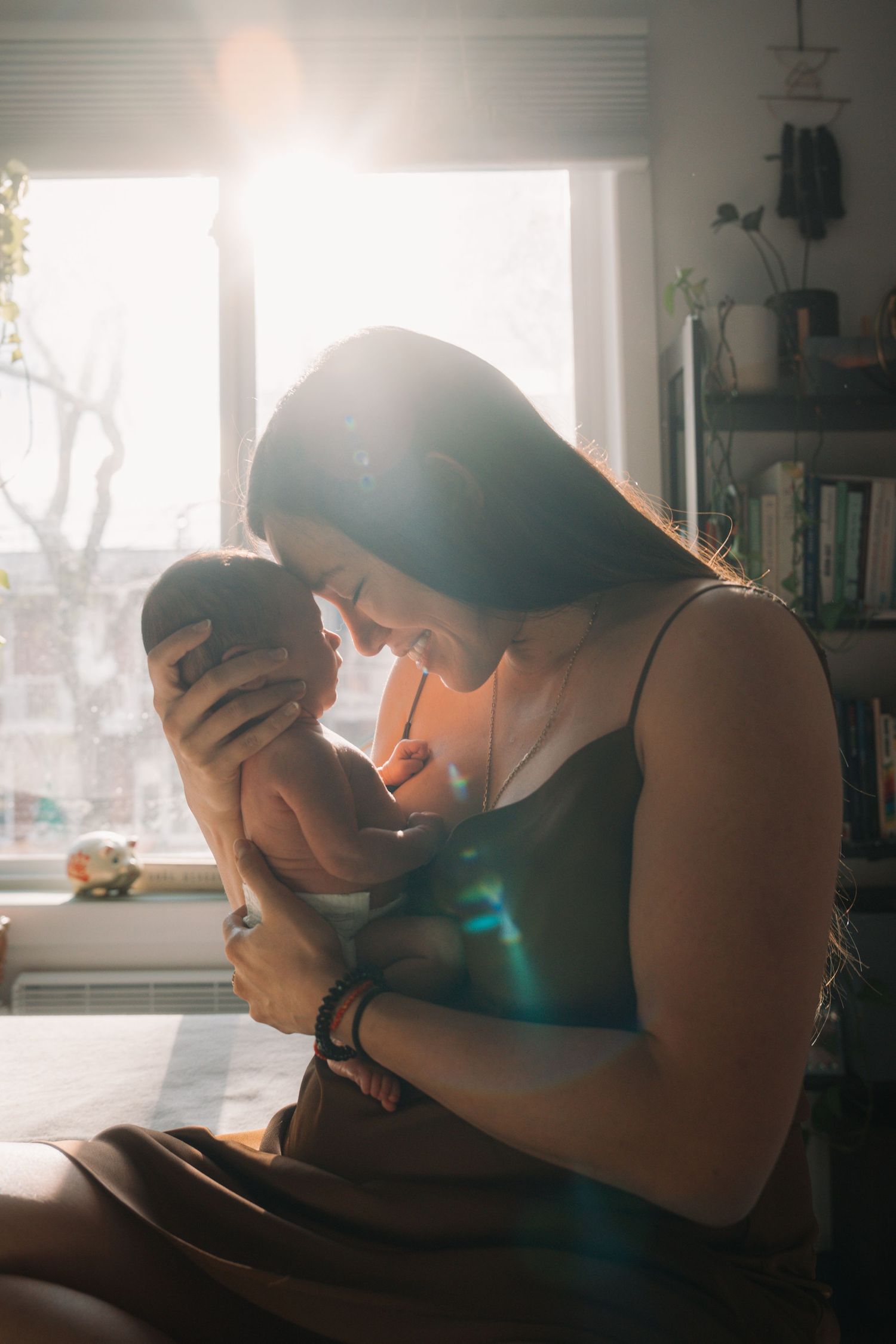 Mother holding newborn baby near sunlit window with beautiful lens flare in cozy home setting.