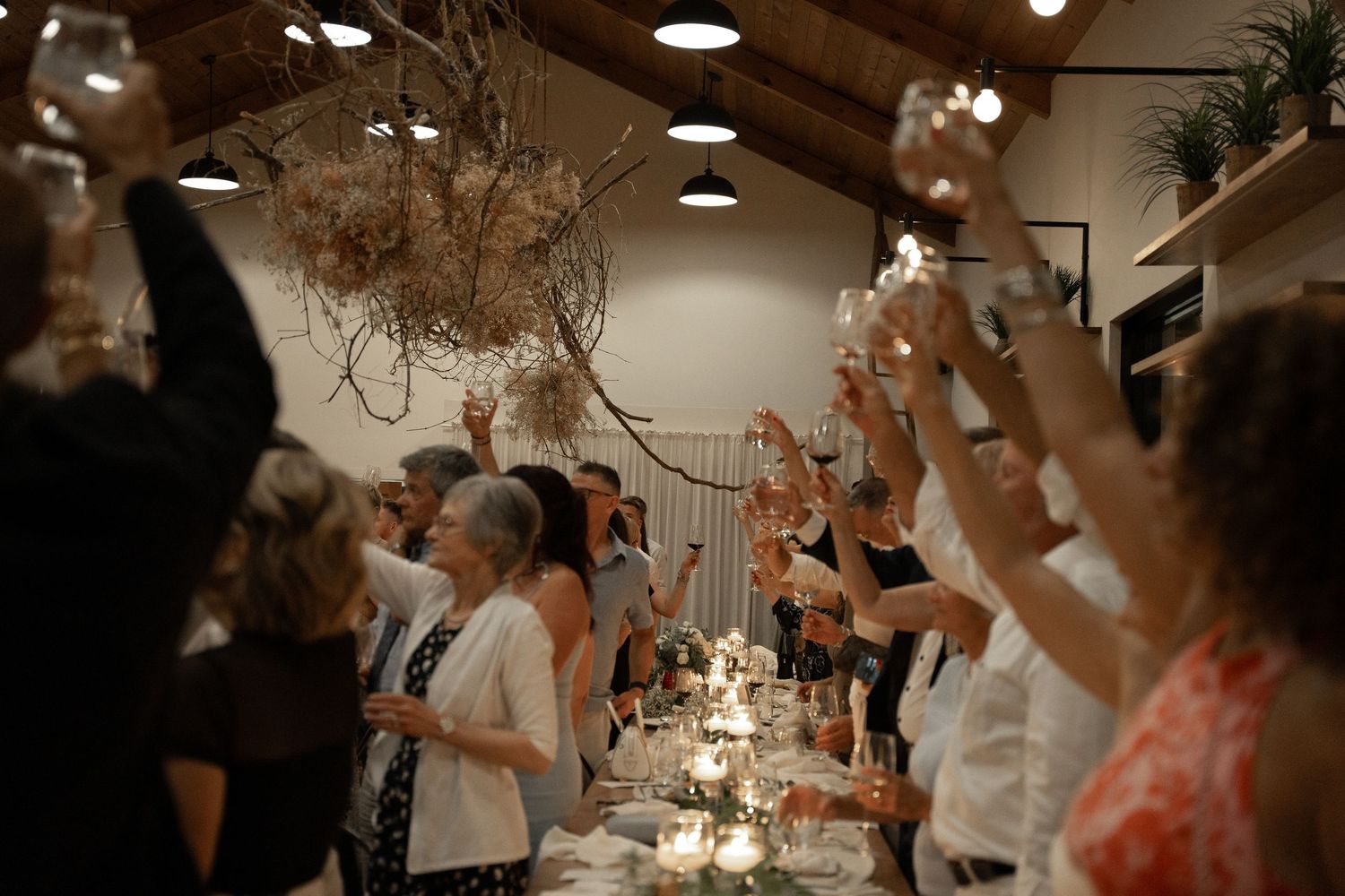 Wedding guests raise glasses in a celebratory toast inside a warmly lit reception venue.