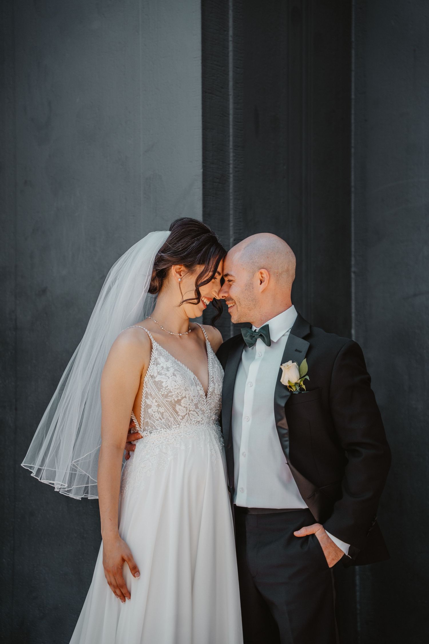 Bride and groom touch foreheads tenderly against a dark gray wall outdoors.