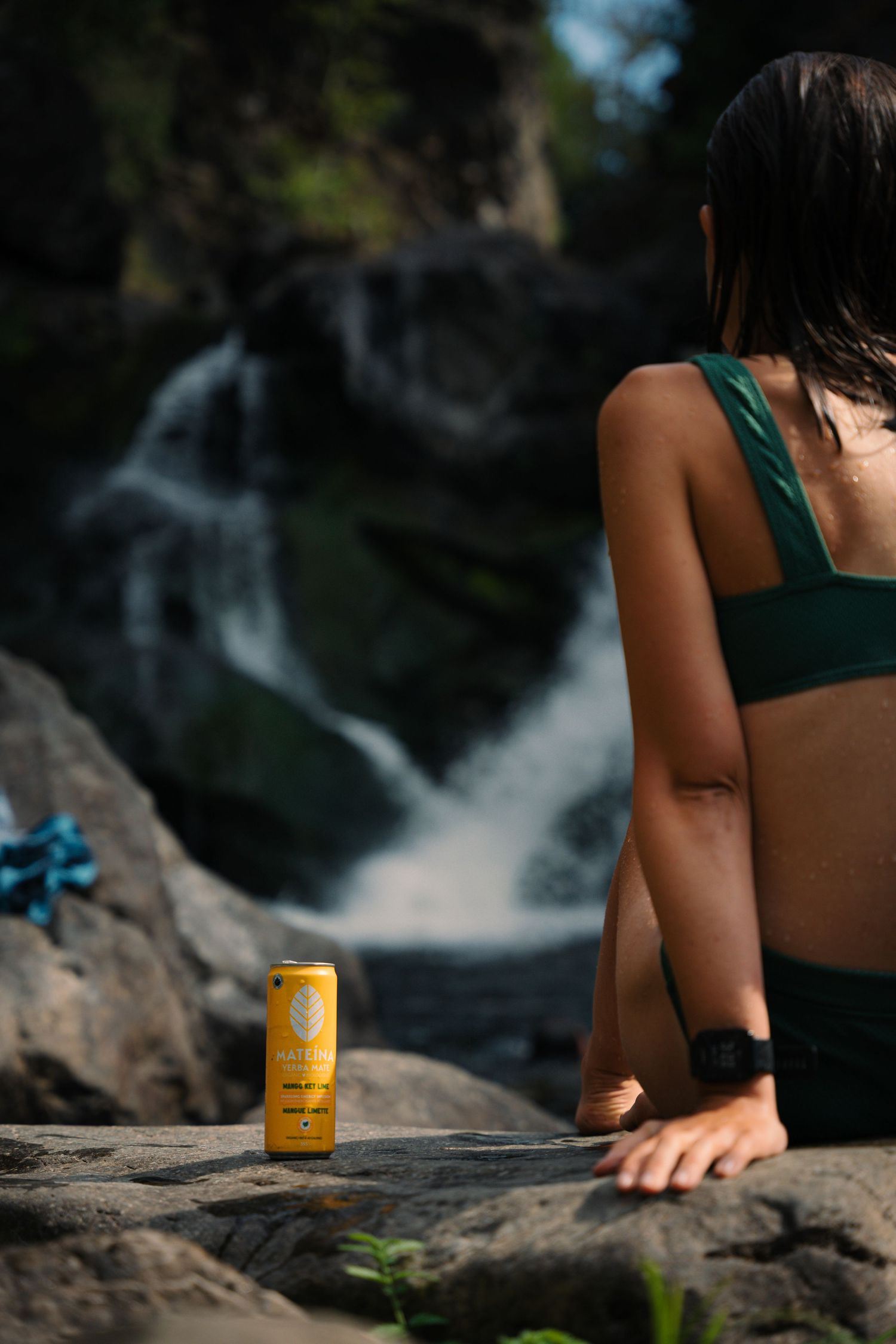 Woman in green bikini top sitting on rocks near waterfall with yellow drink can beside her.
