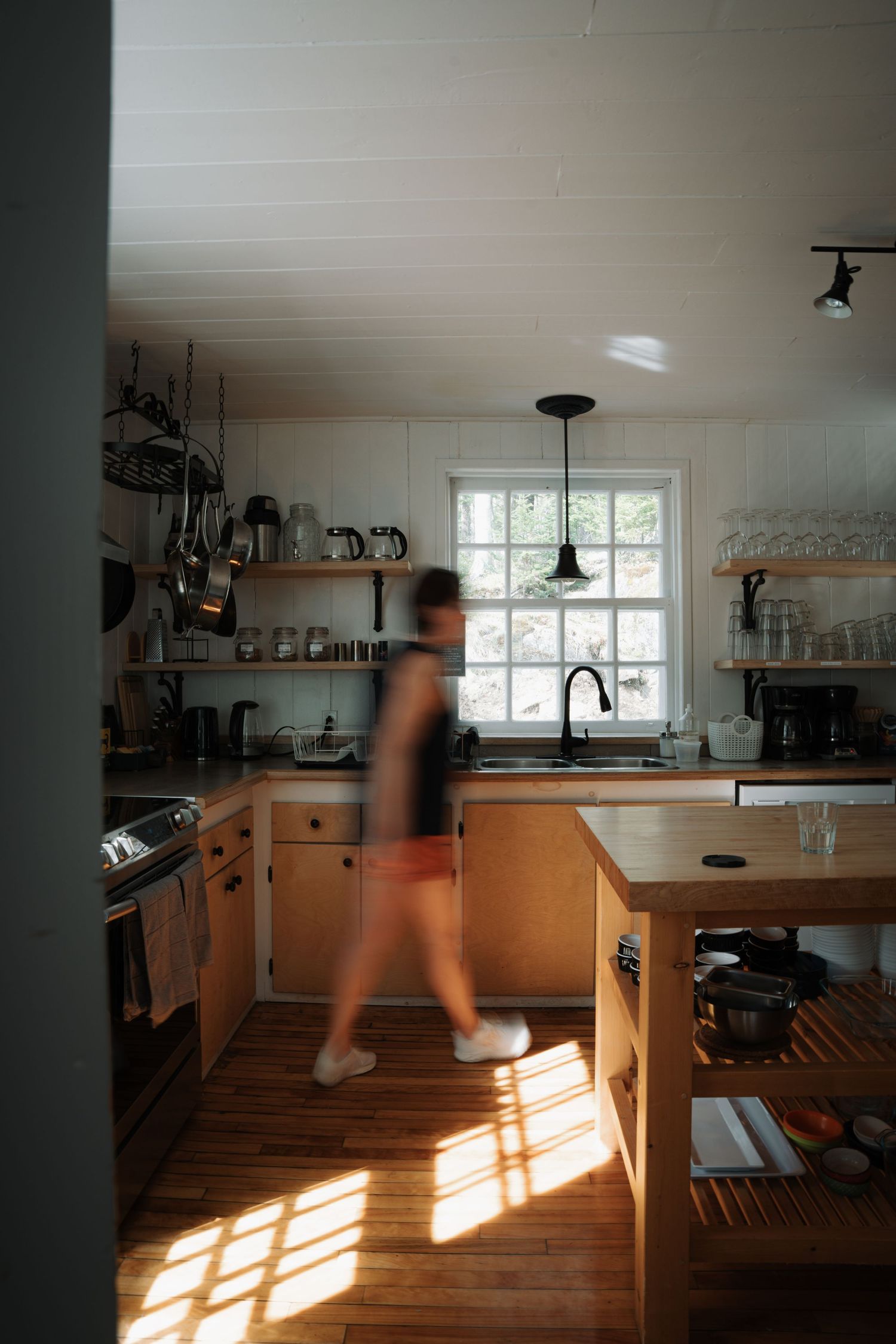 Person walking through rustic kitchen with wooden cabinets and sunlight streaming through window.