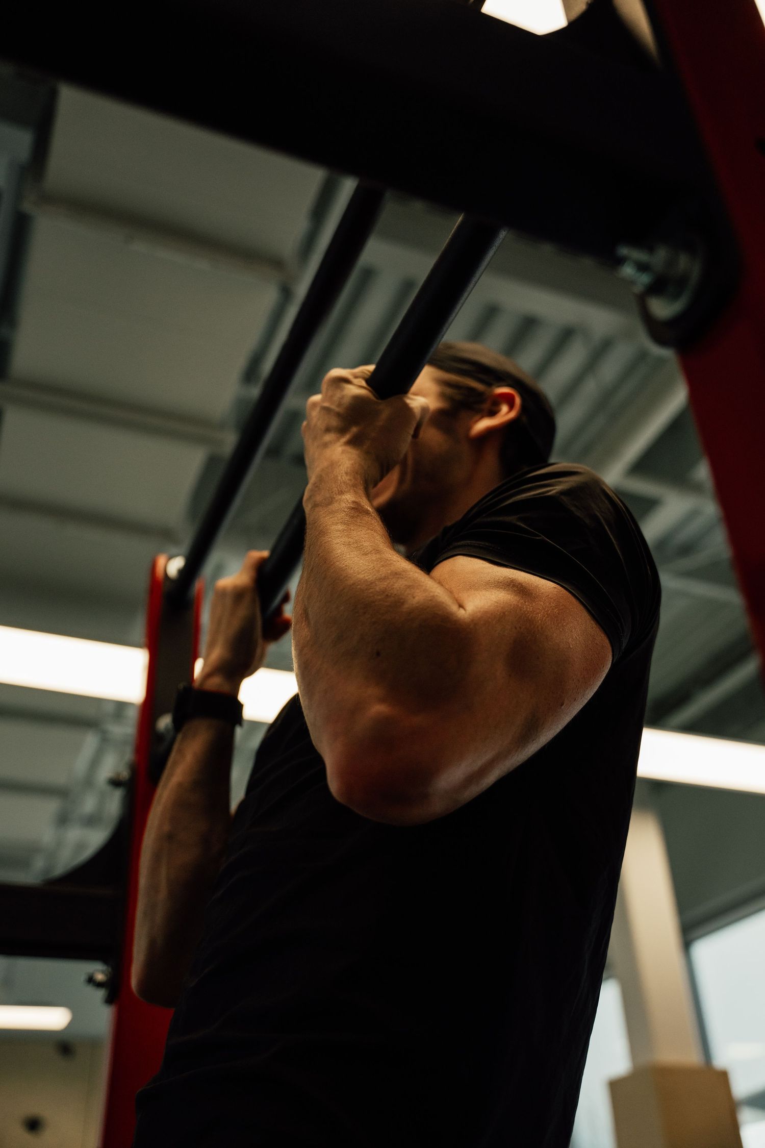 Muscular man in black shirt doing pull-ups on a bar in a gym, showcasing arm strength.
