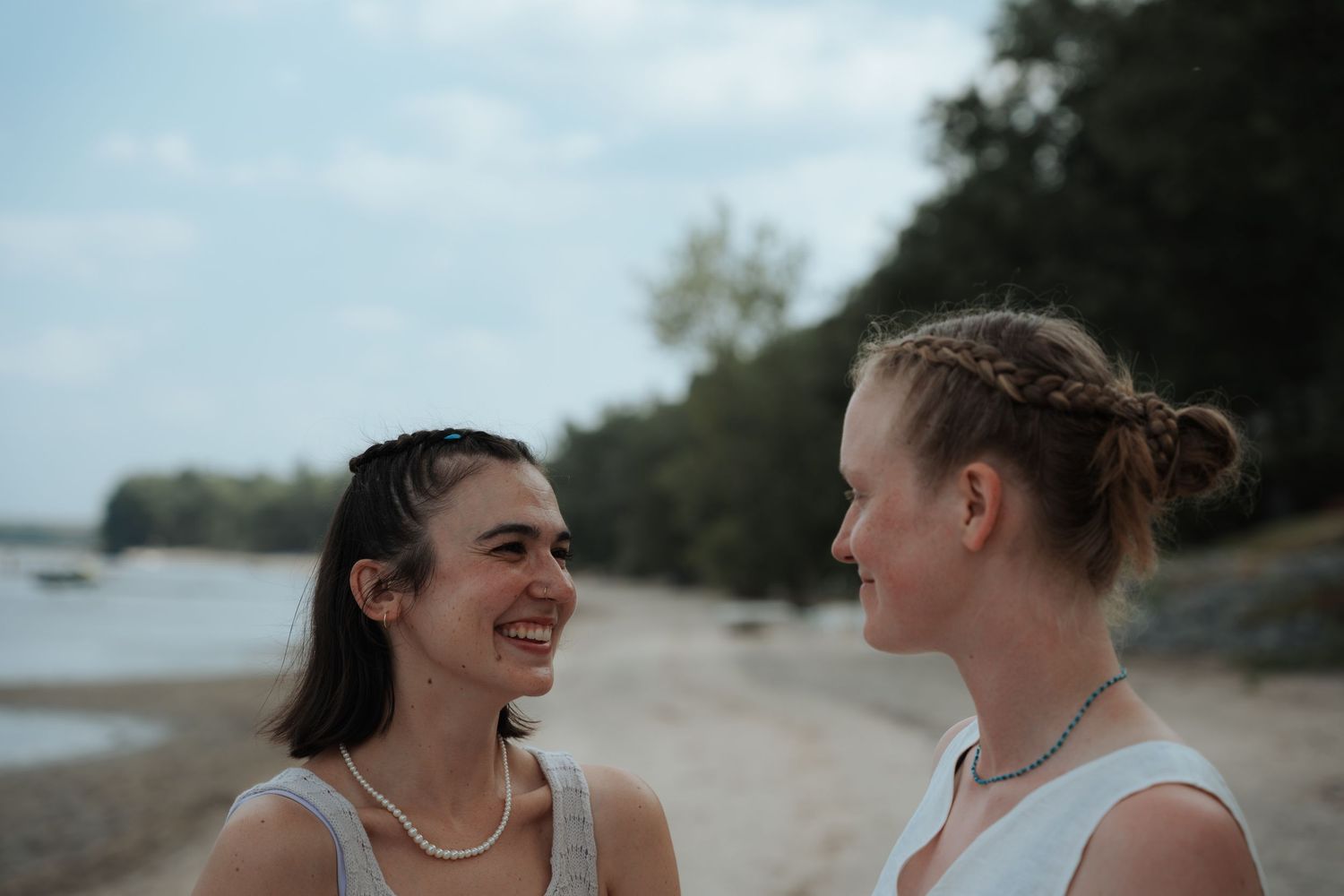 Two brides smiling at each other on a sandy beach during their wedding ceremony.