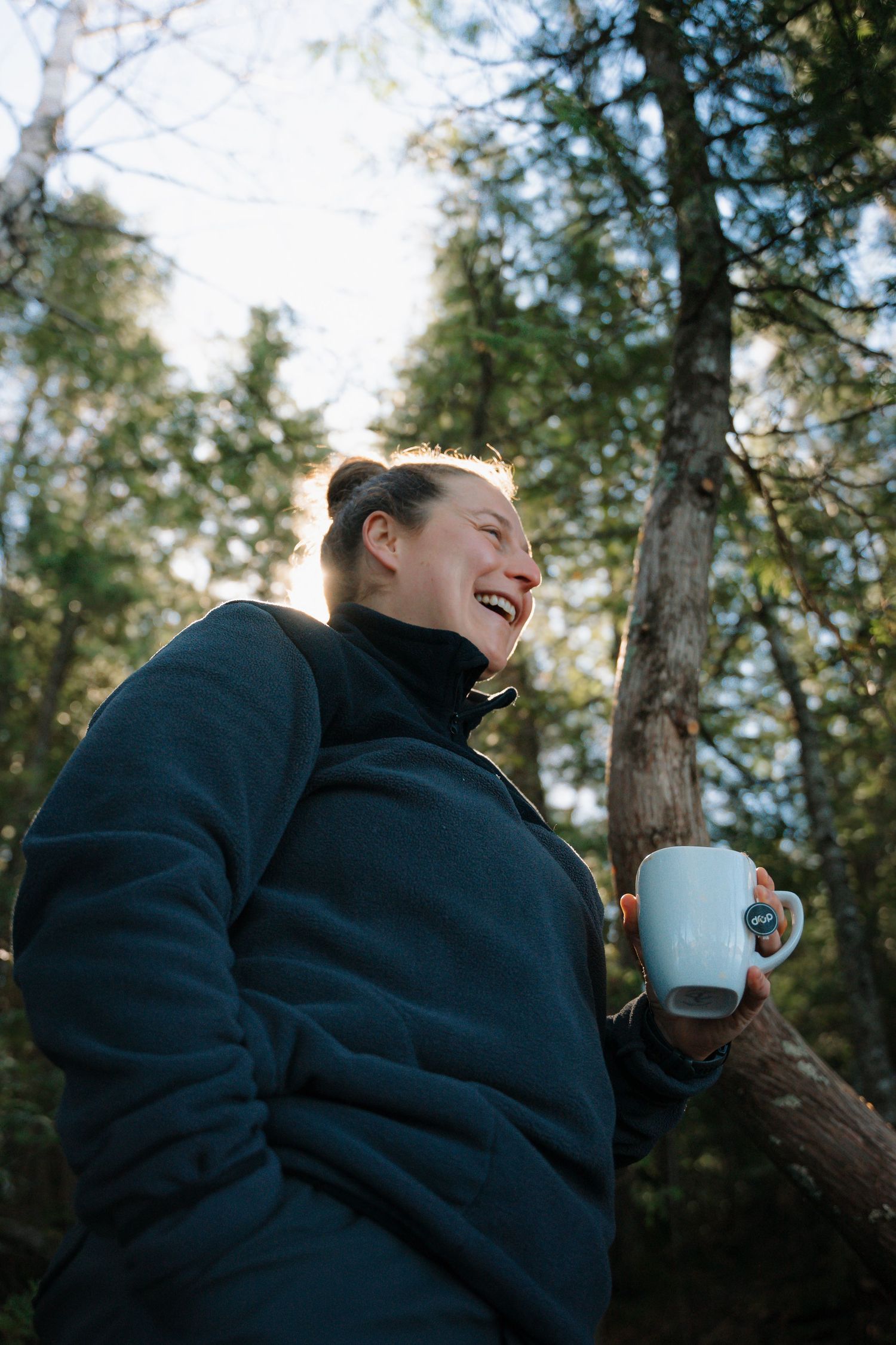 A laughing woman in a dark fleece jacket holds a white mug while standing in a sunlit forest.