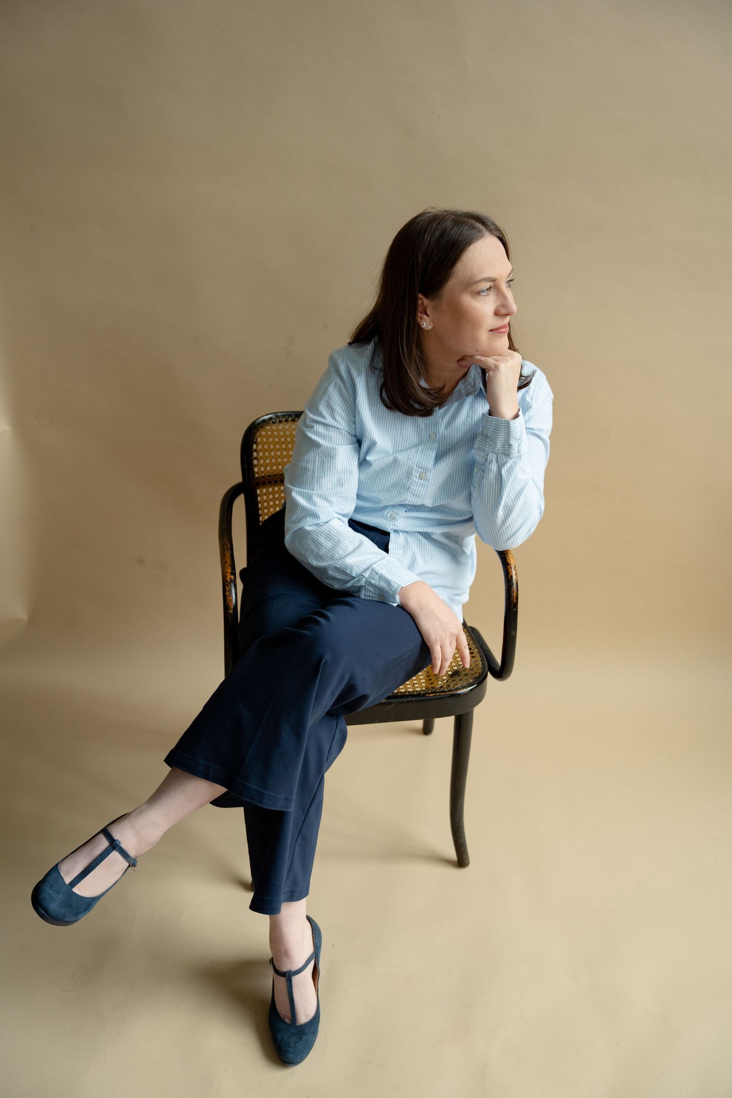 Woman in blue shirt and navy trousers sitting on wooden chair against beige background.