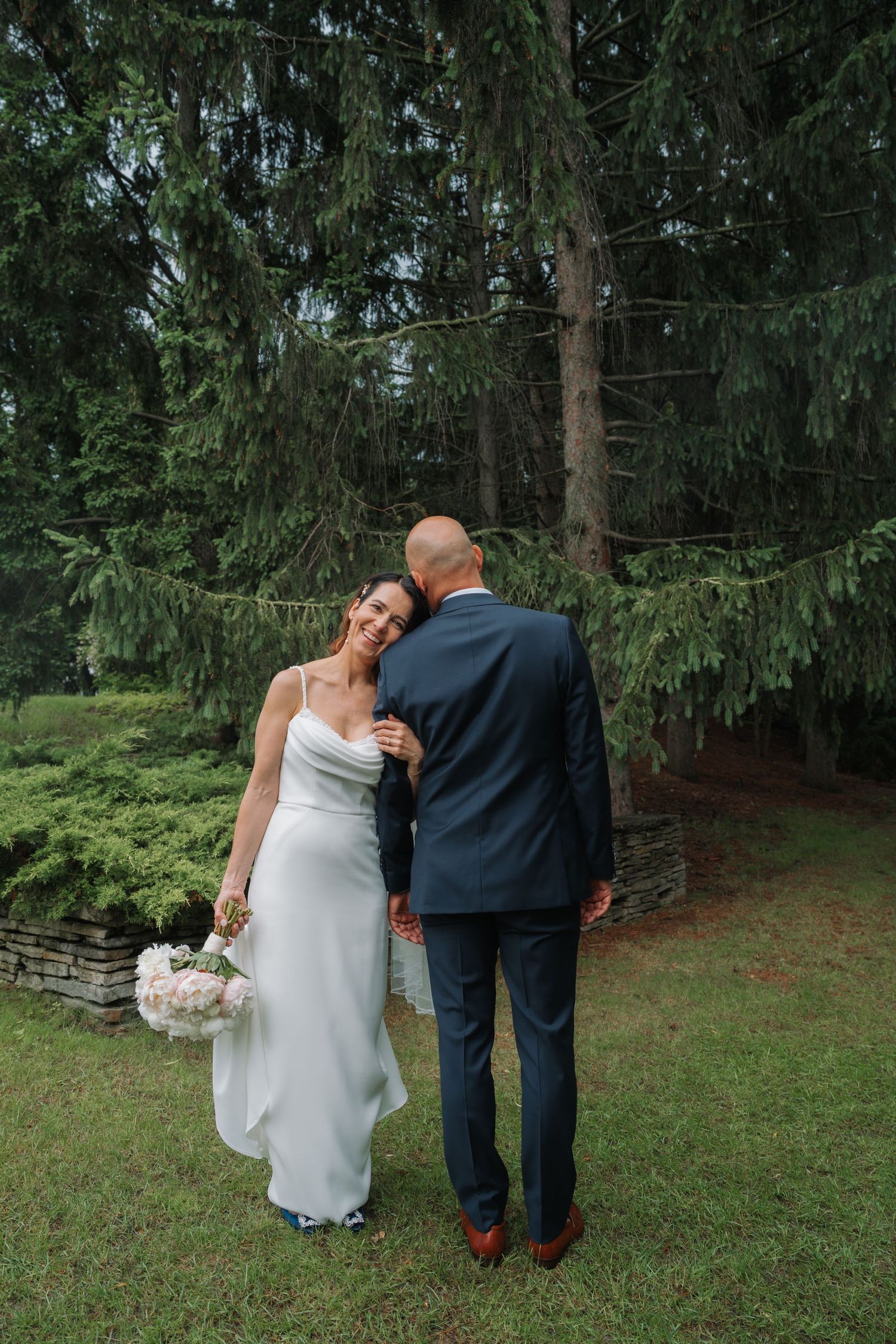 Bride in white gown holds pink bouquet, smiling as groom in navy suit nuzzles her shoulder outdoors.