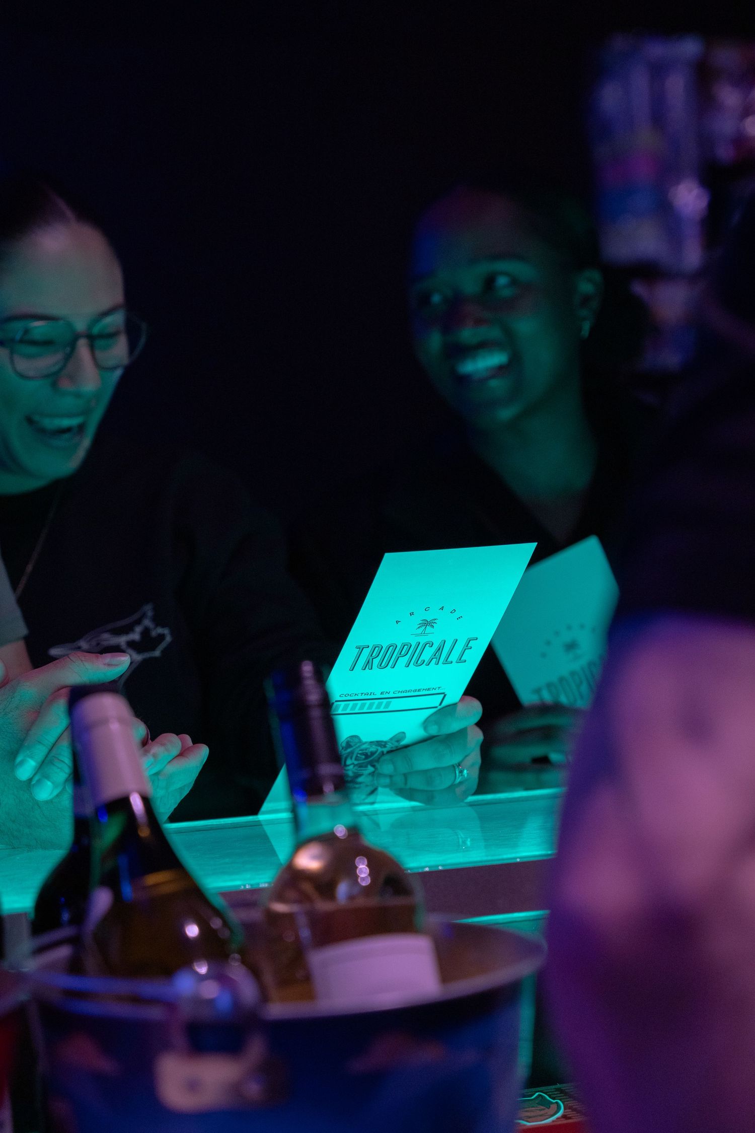 Two people laugh at a bar counter with a Tropicale menu card glowing under neon lights.