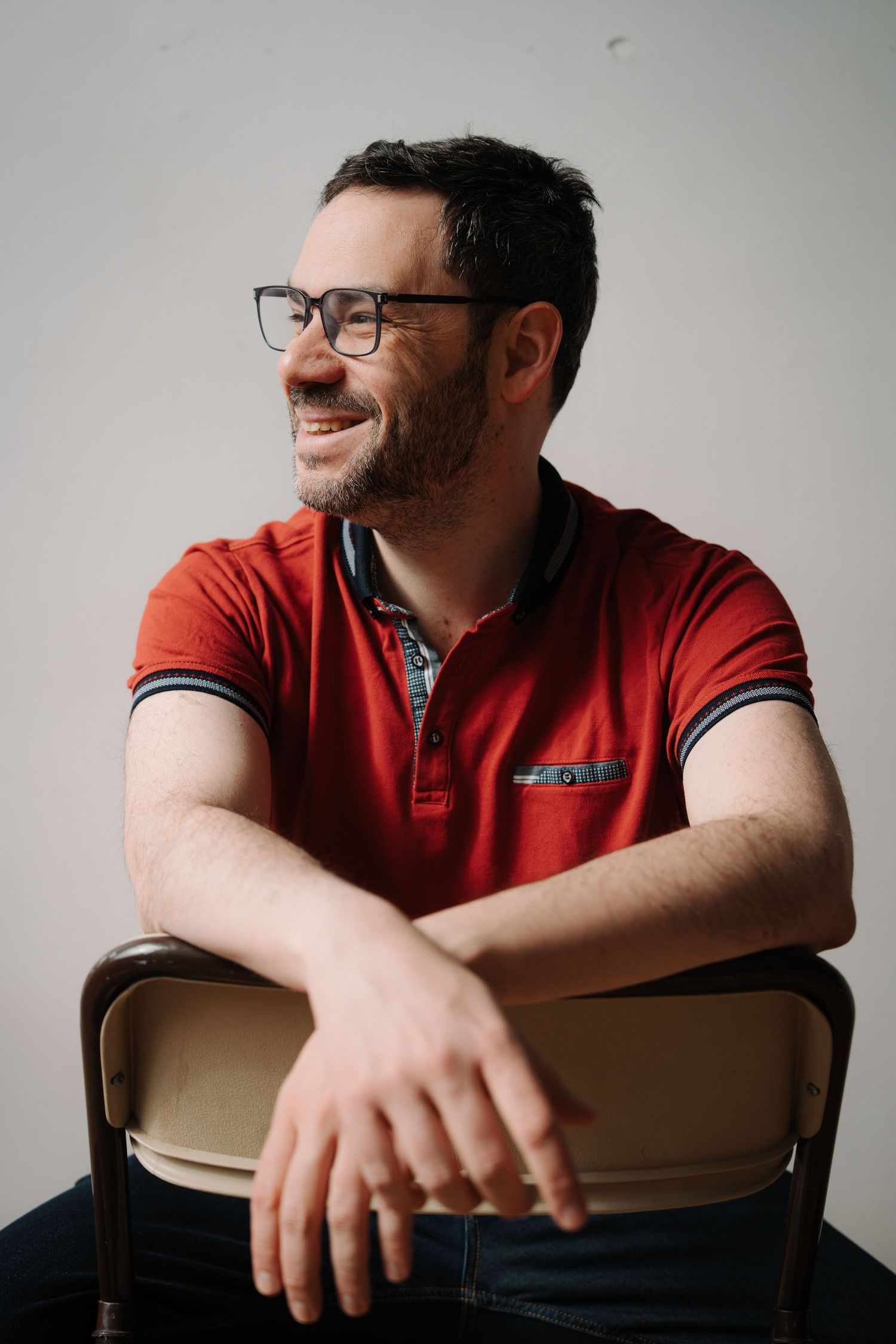 Bearded man in red polo shirt wearing glasses sitting on chair smiling.