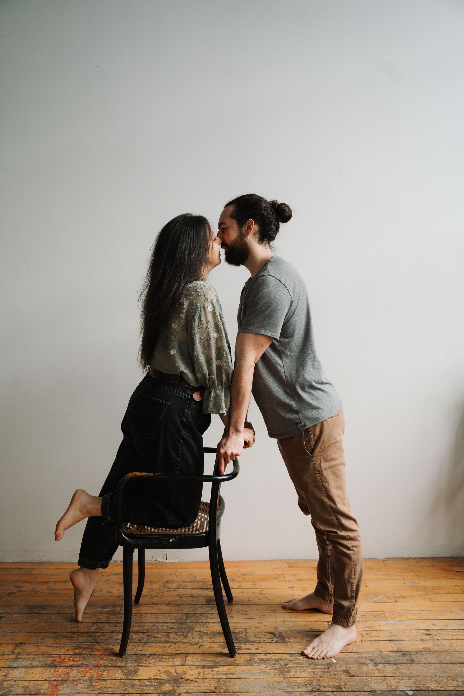 Barefoot couple sharing a kiss while woman stands on a chair in a minimalist studio space.