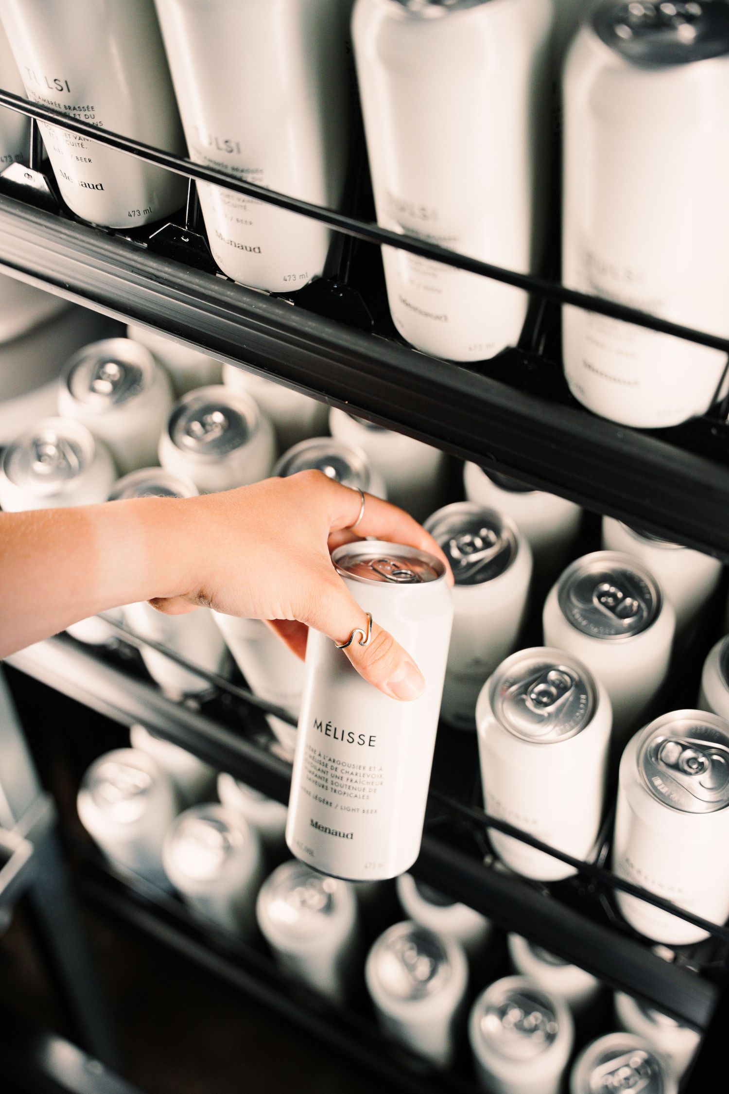 Hand reaching into a refrigerator to grab a white can of Melisse sparkling beverage from a stocked shelf.
