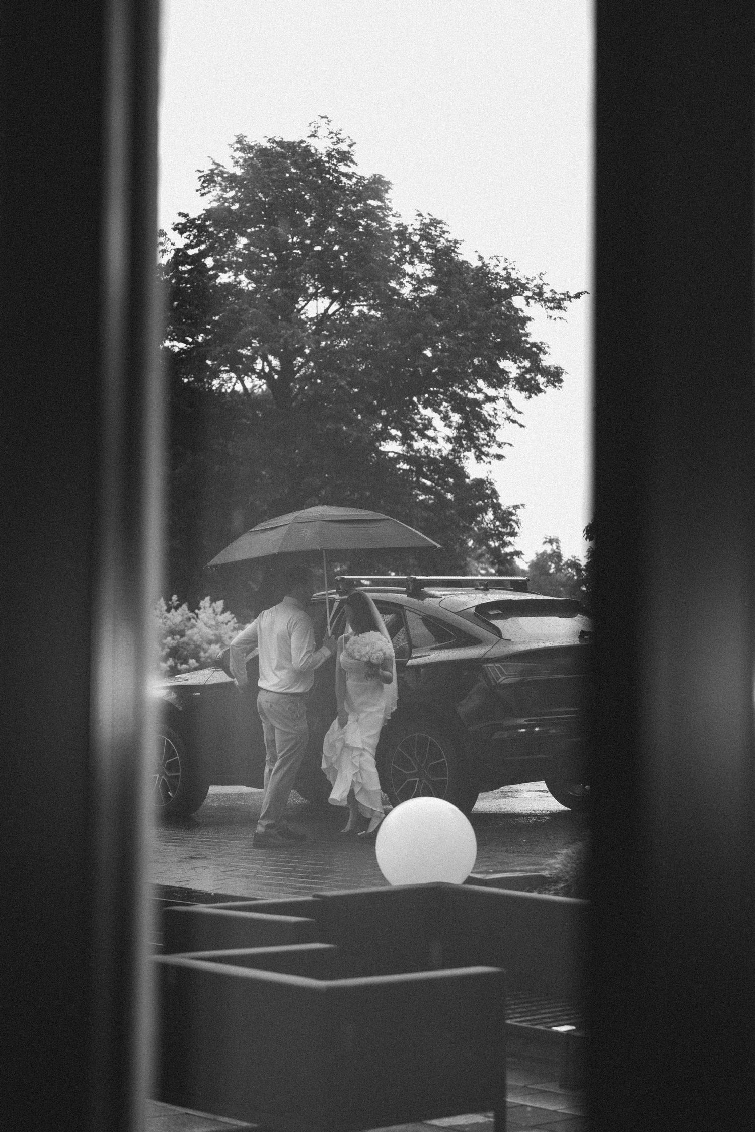 Black and white photo of couple sharing umbrella in rain near car, with trees in background.