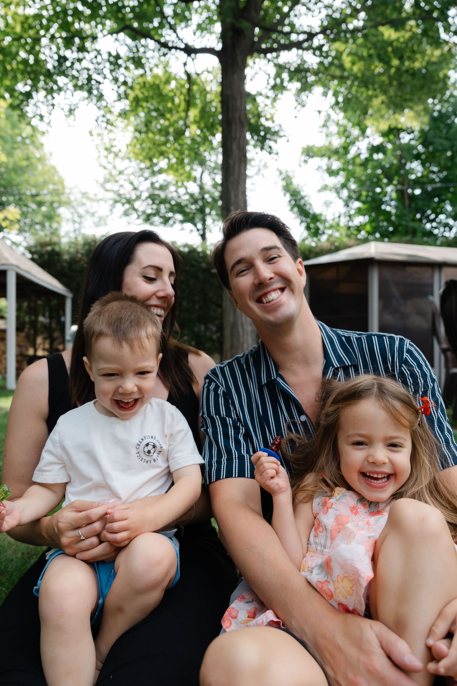 Happy family of four smiling outdoors, parents holding two young children in a lush green backyard setting.