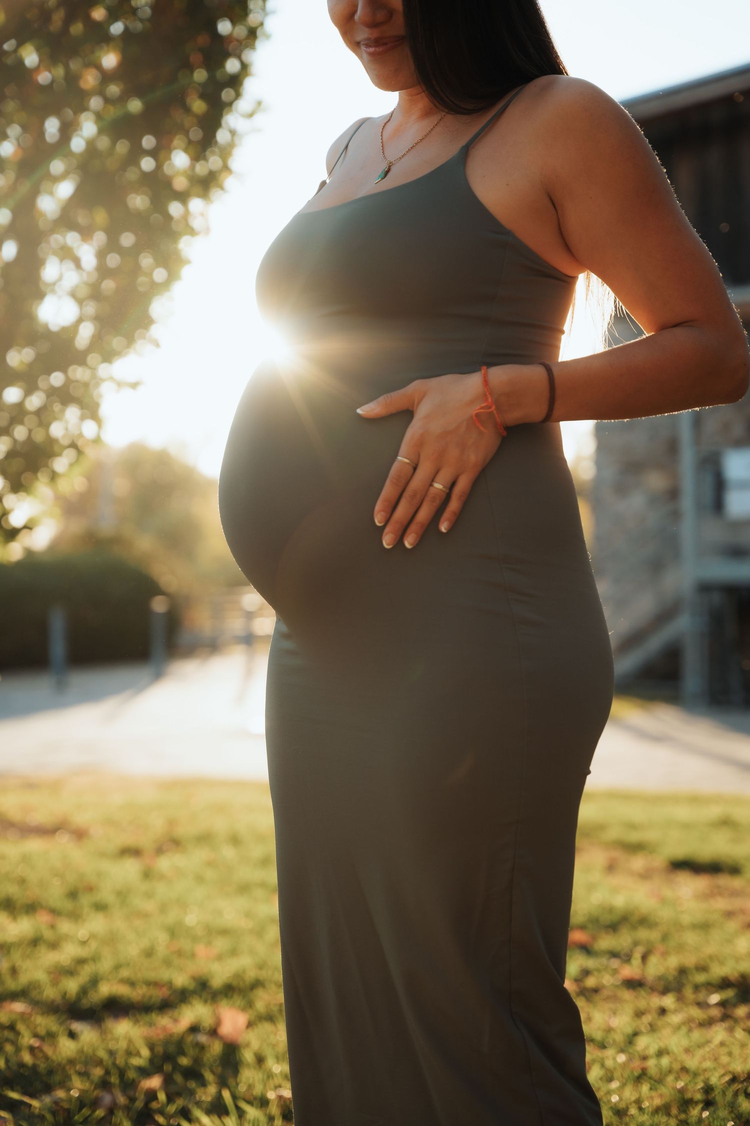 Pregnant woman in olive green maxi dress holding her baby bump outdoors with sunlight glowing behind her.