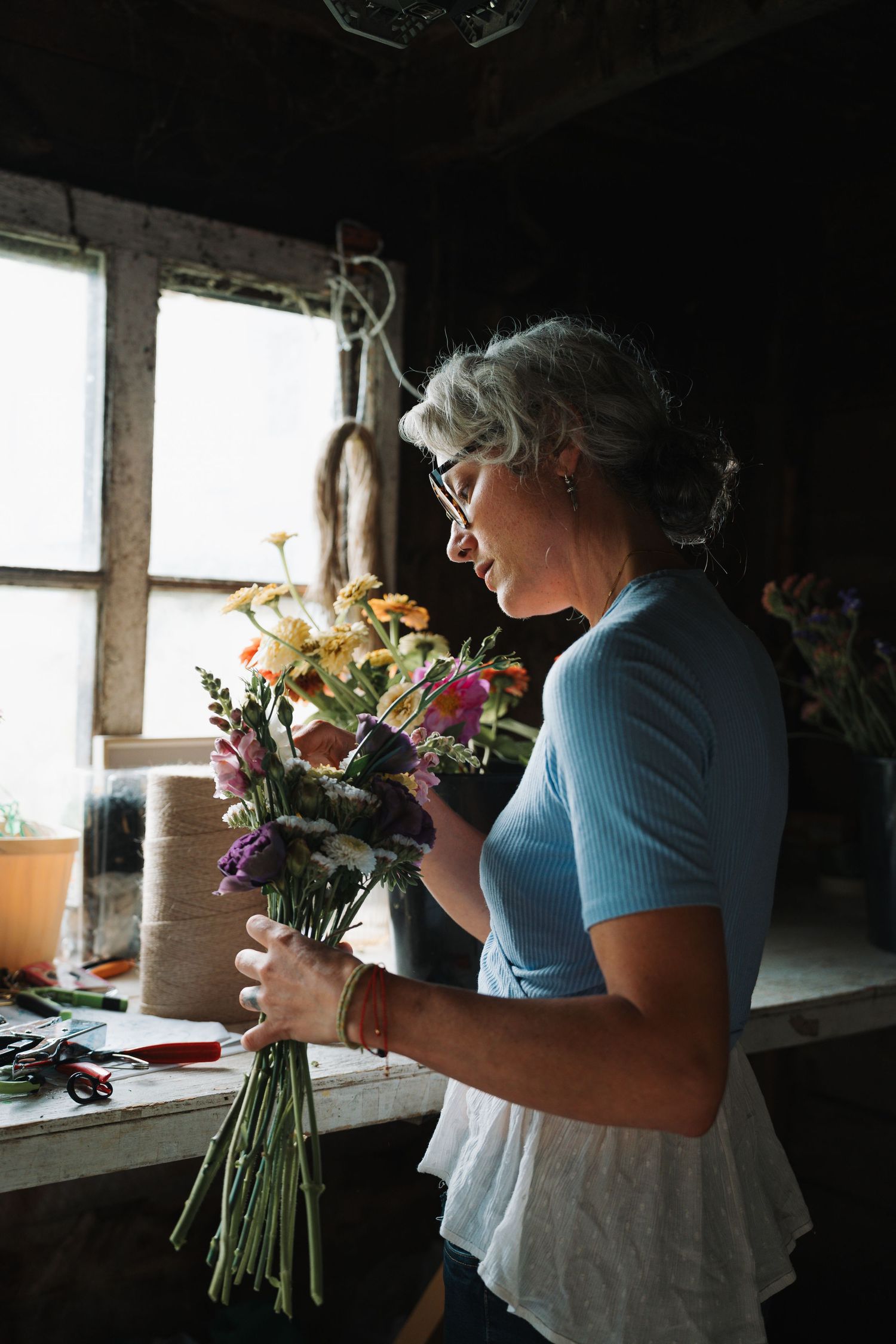 Older woman with gray hair arranging a colorful bouquet of wildflowers in a rustic floral workshop.