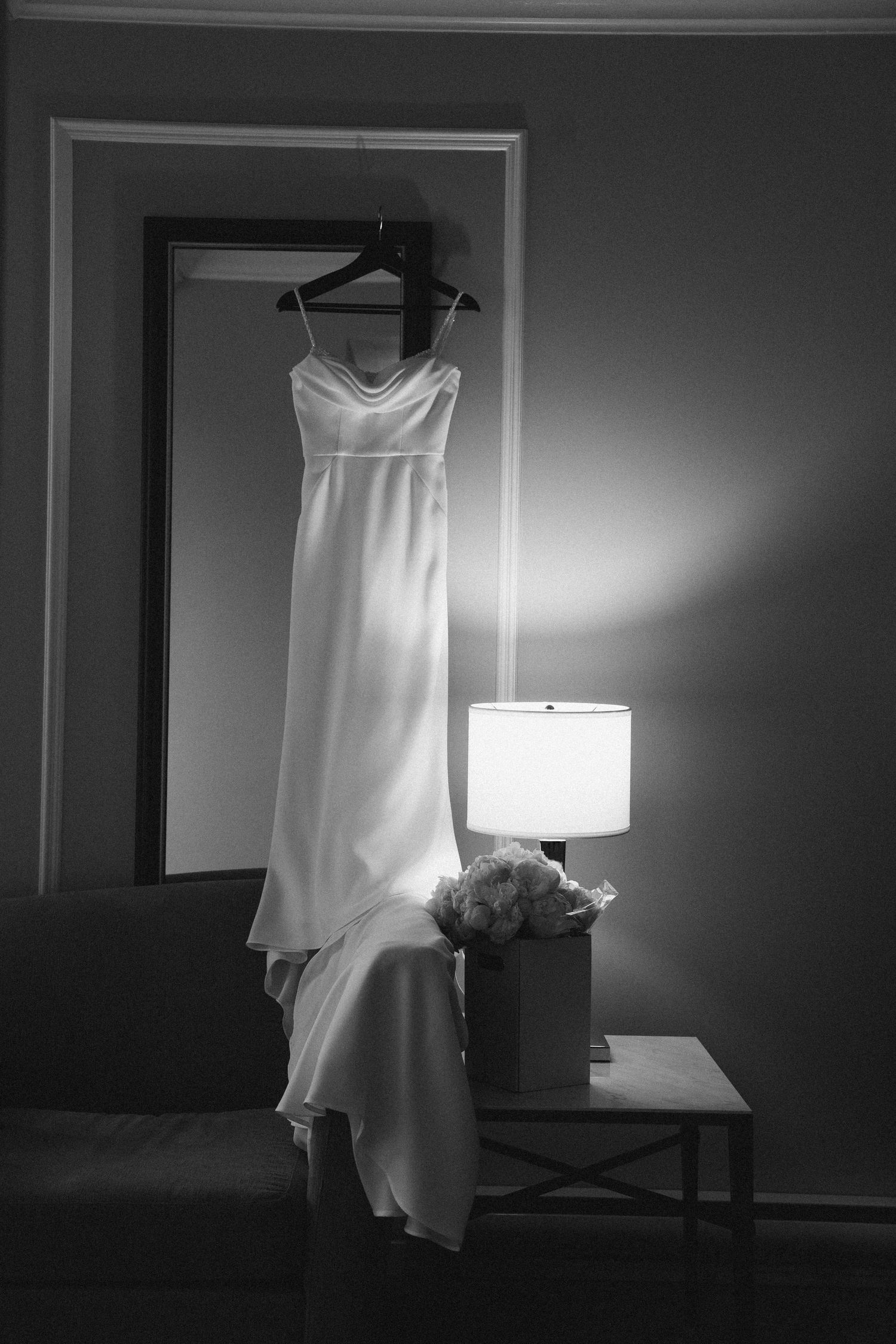 Black and white photo of an elegant wedding dress hanging near a glowing lamp with a bridal bouquet on a side table.
