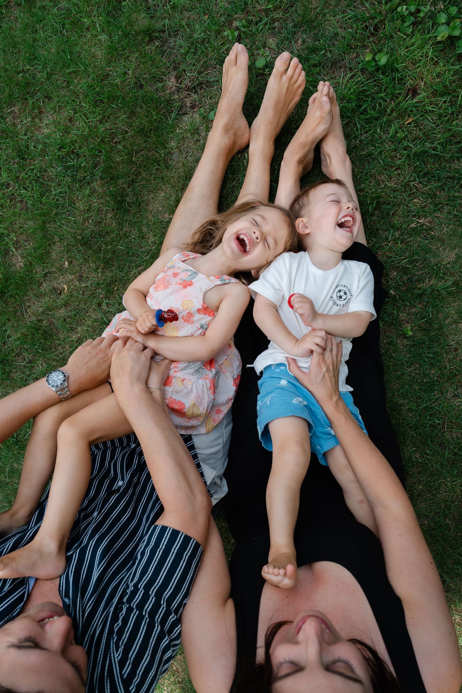Family laughing together on grass, two adults lying down with children resting on their stomachs outdoors.