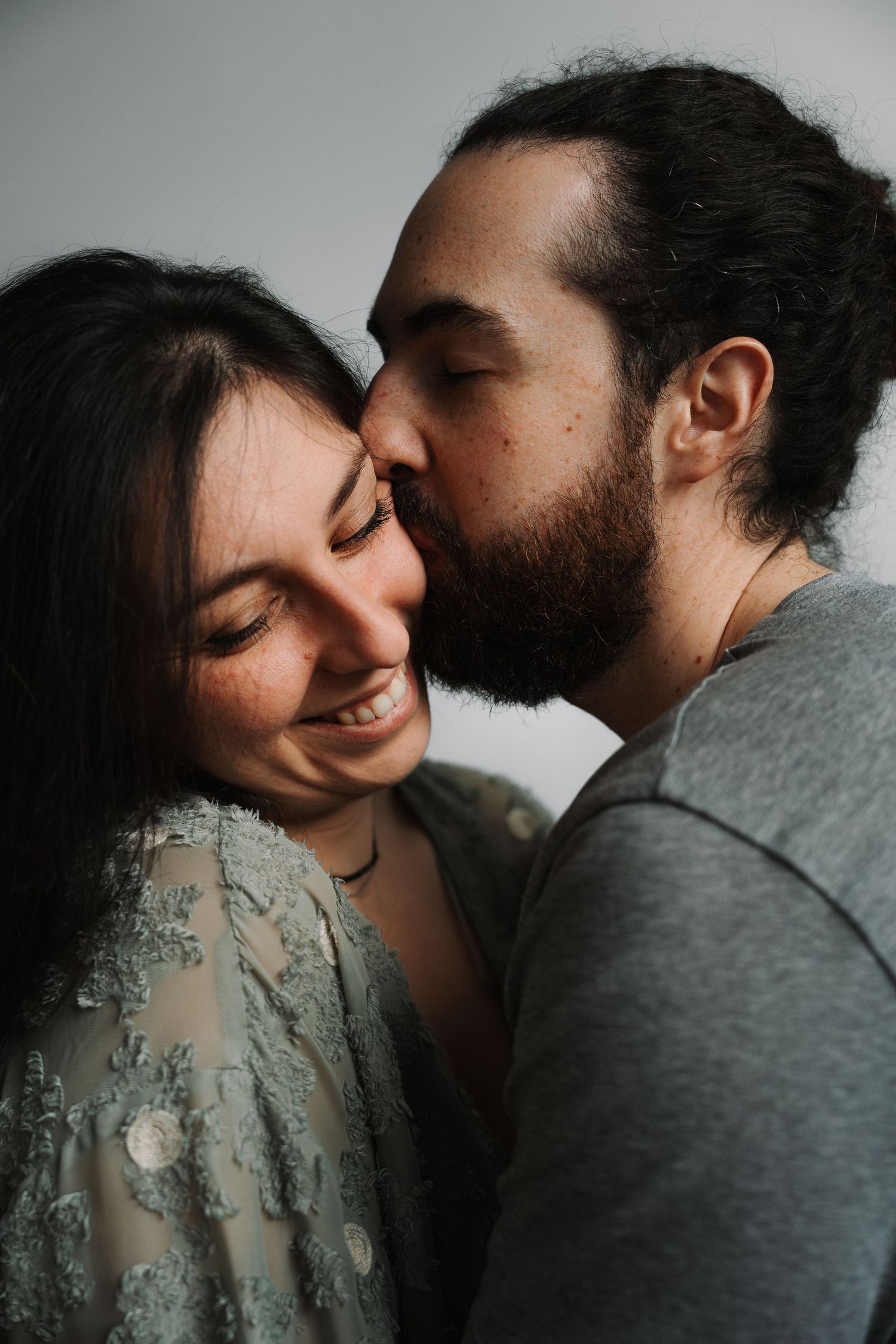 Bearded man with hair bun gently kissing smiling dark-haired woman on the cheek indoors.