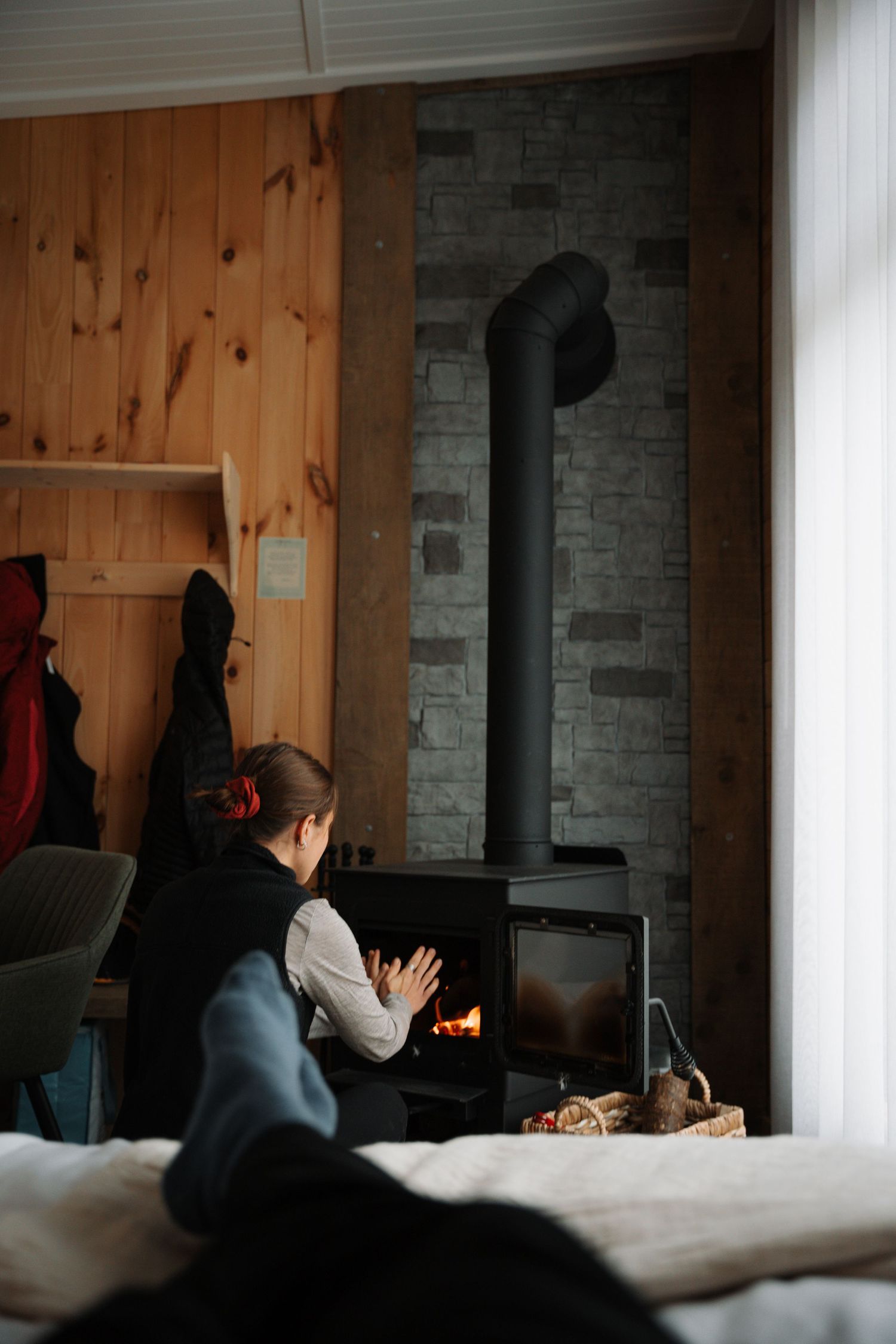 Child warming hands by black wood burning stove in cozy wooden cabin interior.