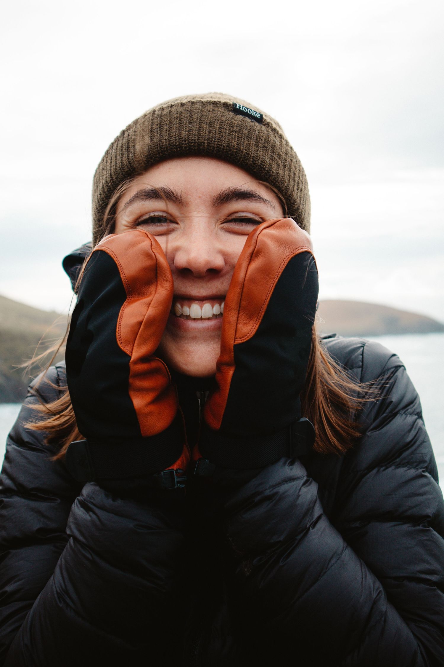 Smiling woman in black puffer jacket and beanie pressing orange mittens to her cheeks outdoors.