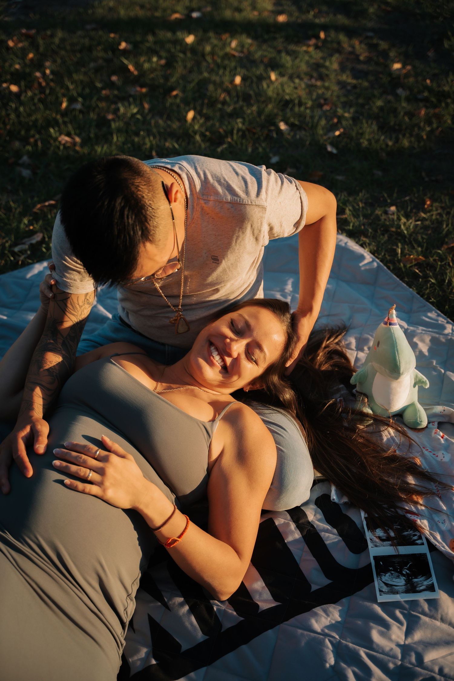 Couple relaxing on a blanket outdoors at golden hour, pregnant woman smiling as partner embraces her.