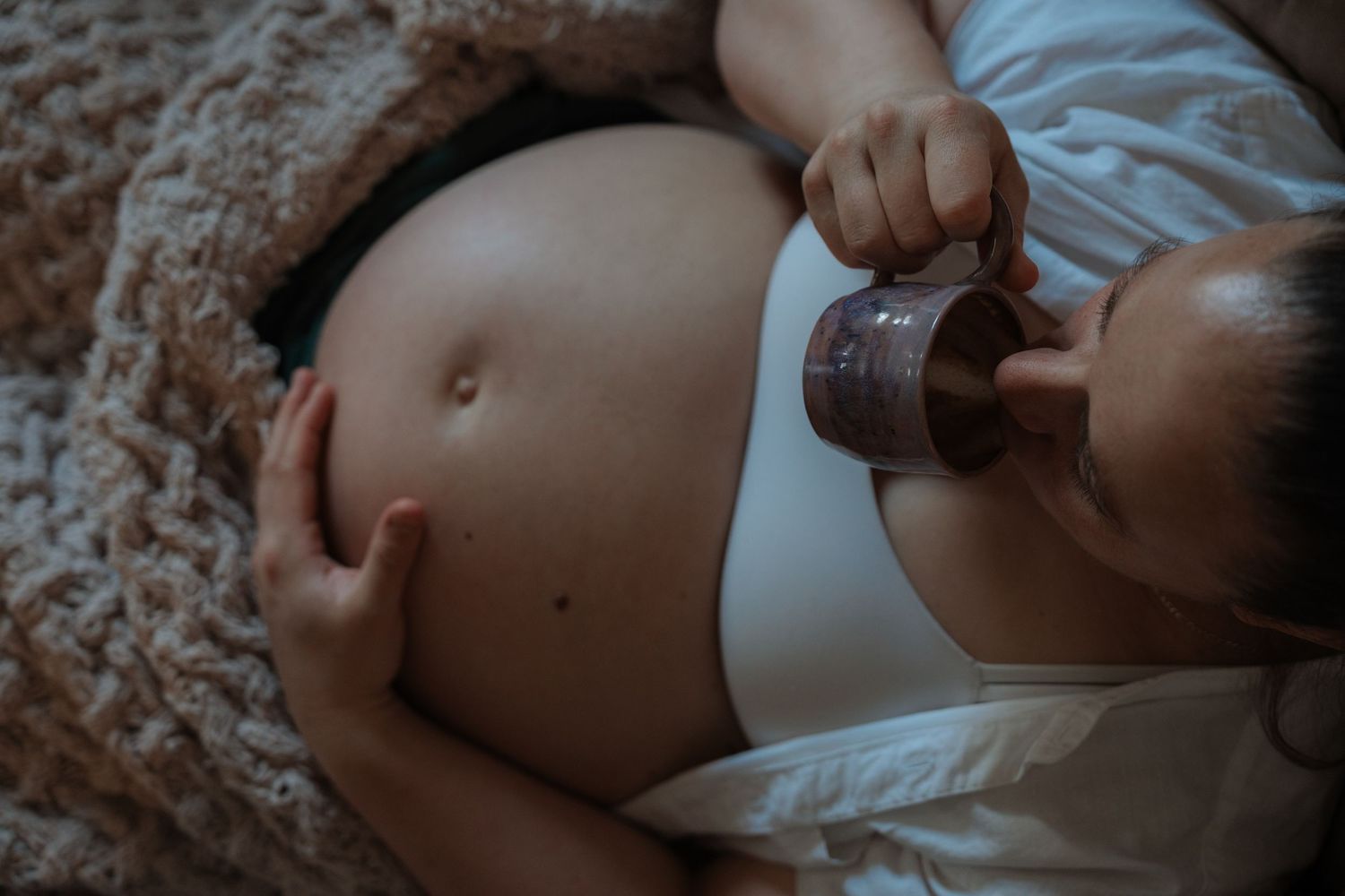 Pregnant woman lying down, cradling her bare baby bump while holding a small cup.