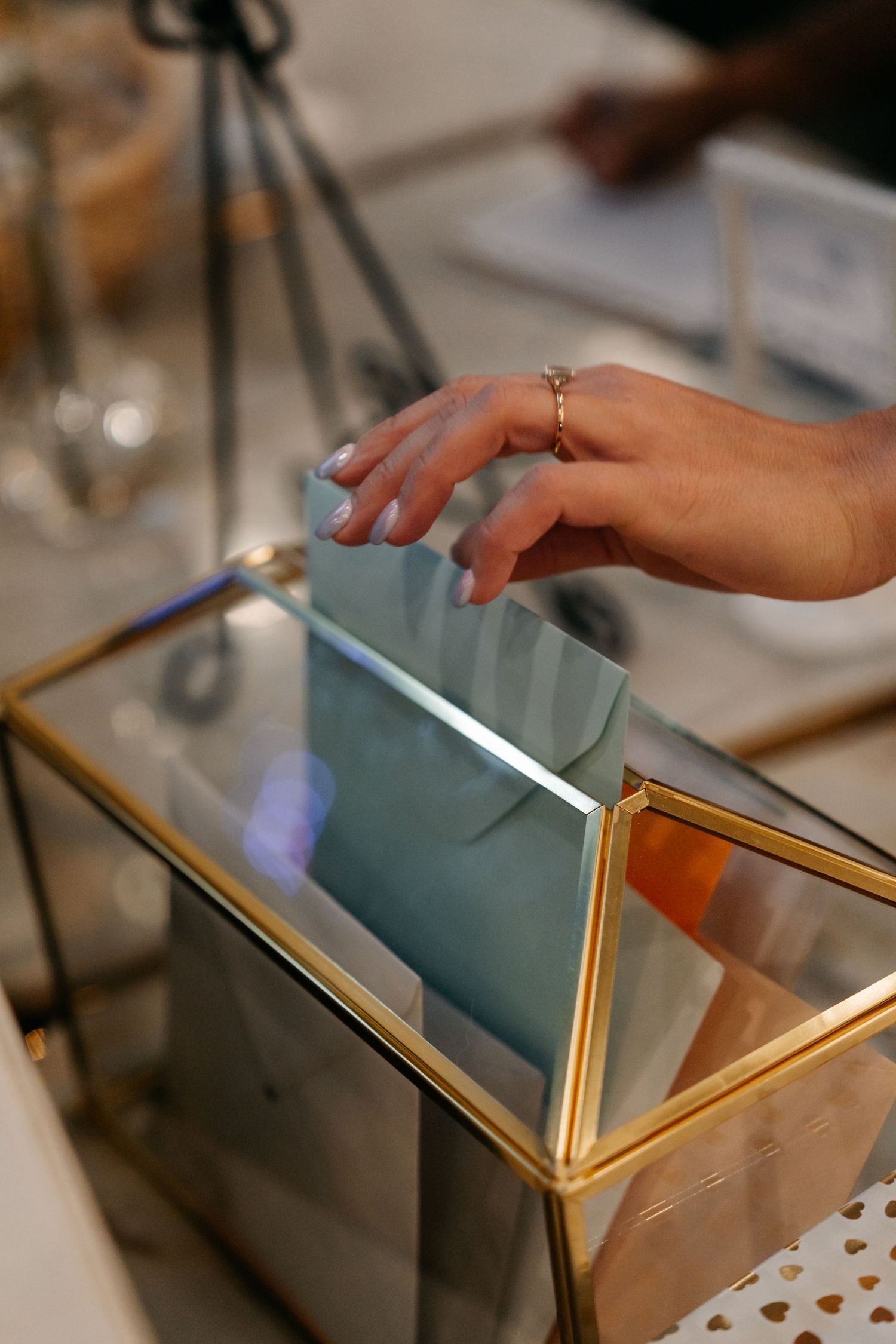 Hand placing light blue envelope into gold and glass wedding card box at reception.