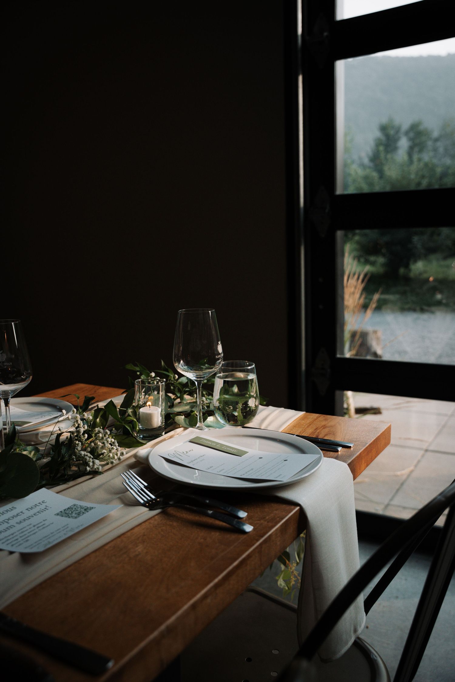 Elegant wedding reception table setting with greenery, wine glasses, and menu cards by a window.