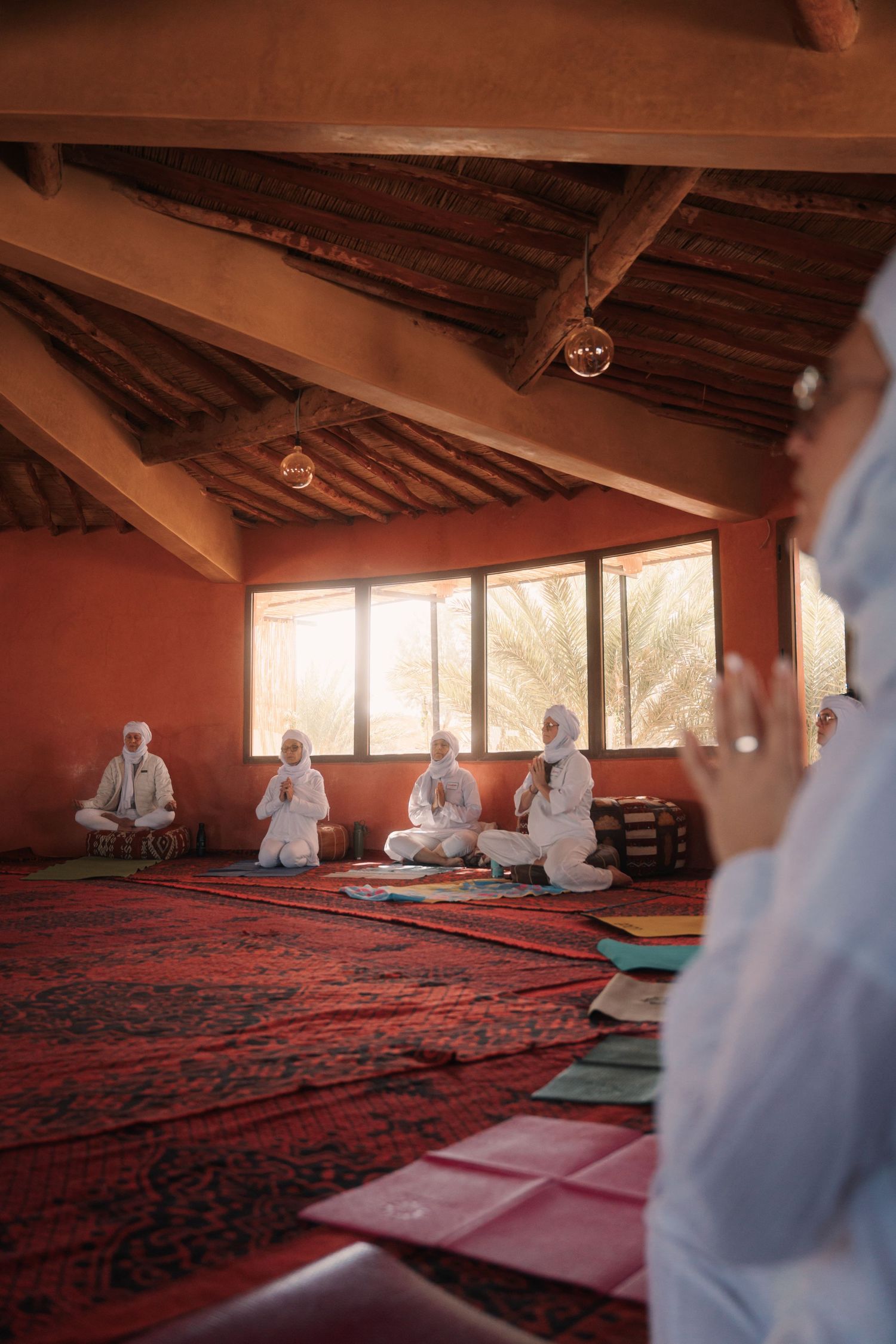 A yoga instructor leads a small group meditation session in a rustic Moroccan desert retreat with red walls.