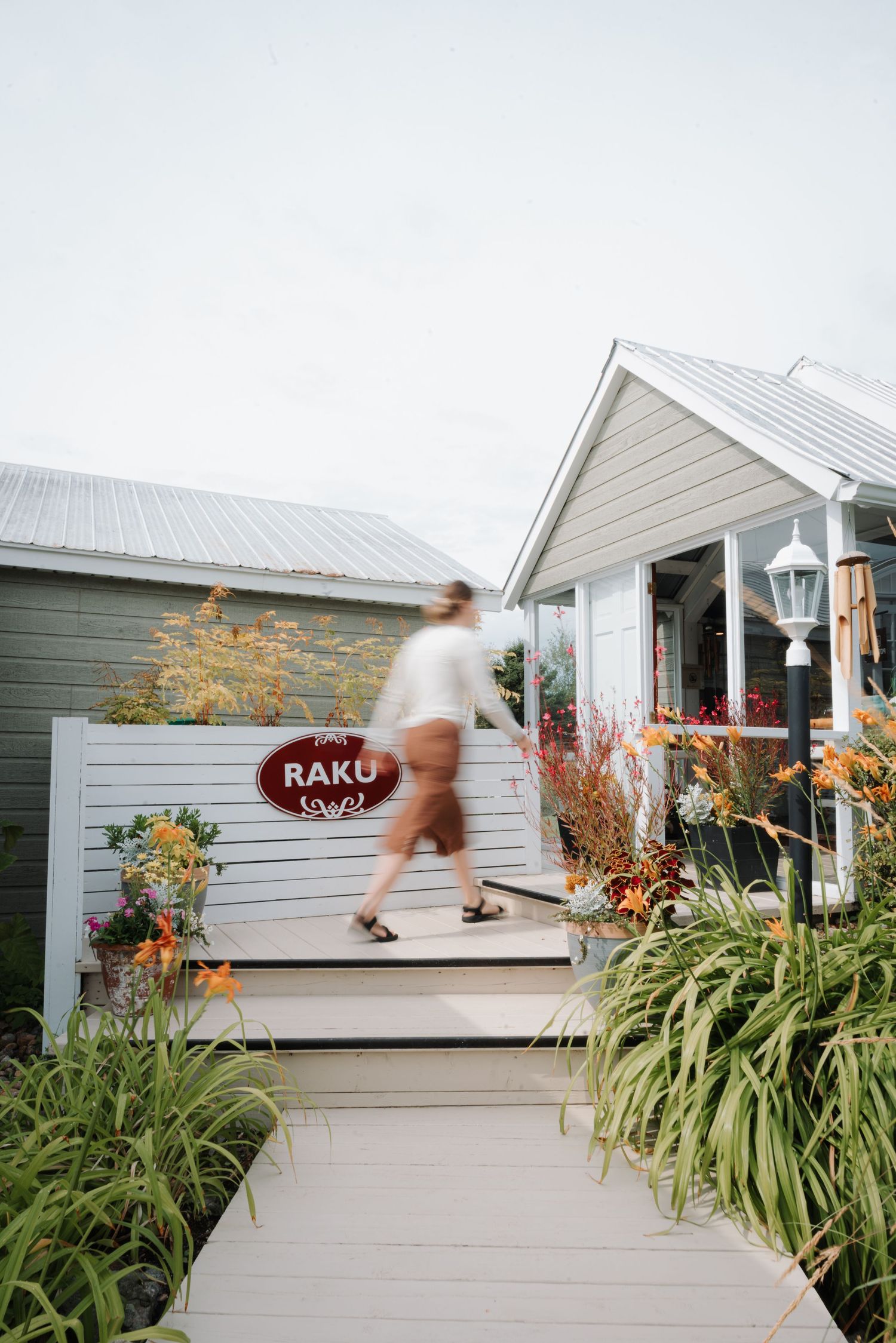 Person walking up wooden steps to a charming gray cottage with a red Raku sign, surrounded by lush garden plants.