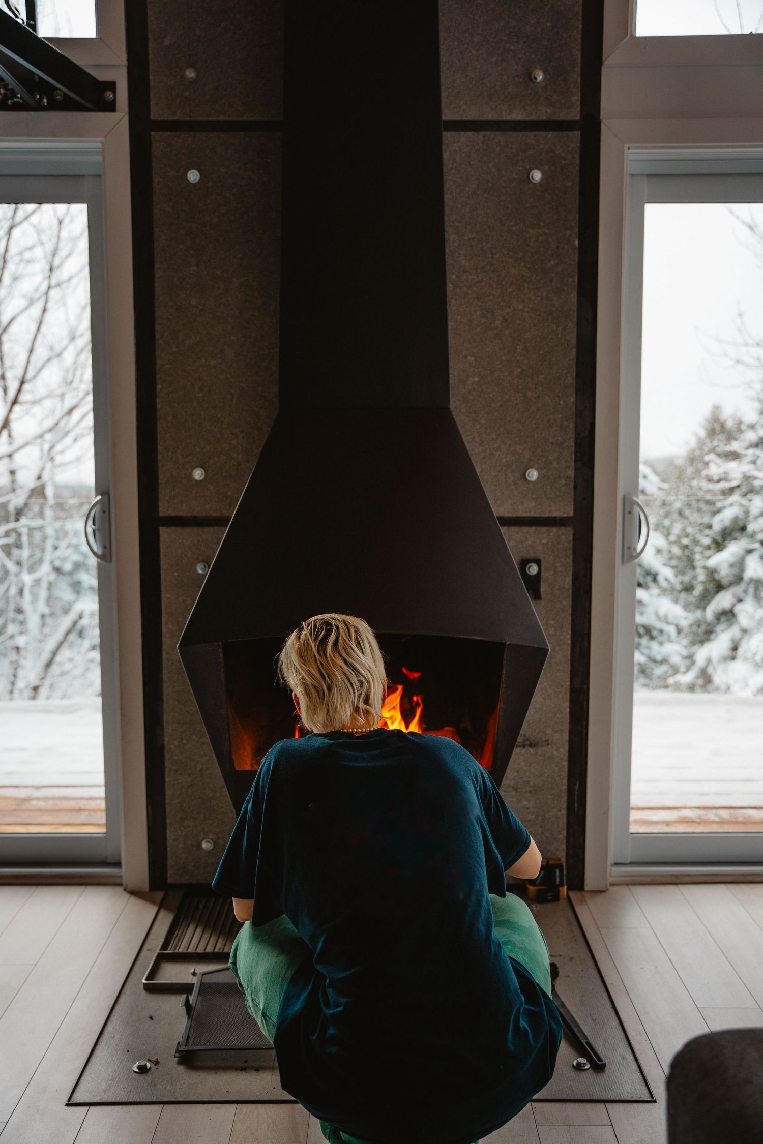Blonde person sitting on floor in front of a modern fireplace with snowy forest view outside.