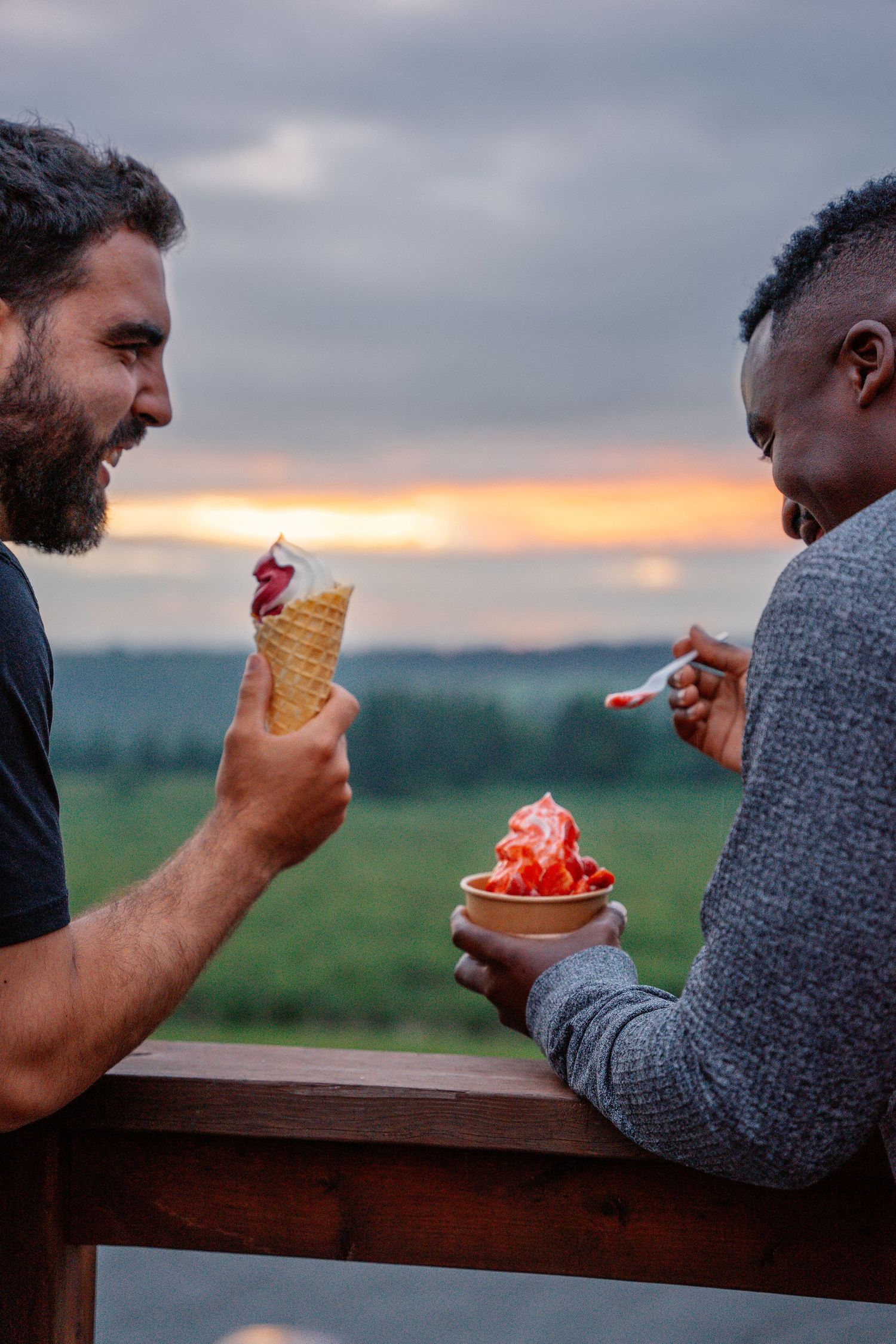 Two men laughing and enjoying ice cream cones together on an outdoor deck at sunset.