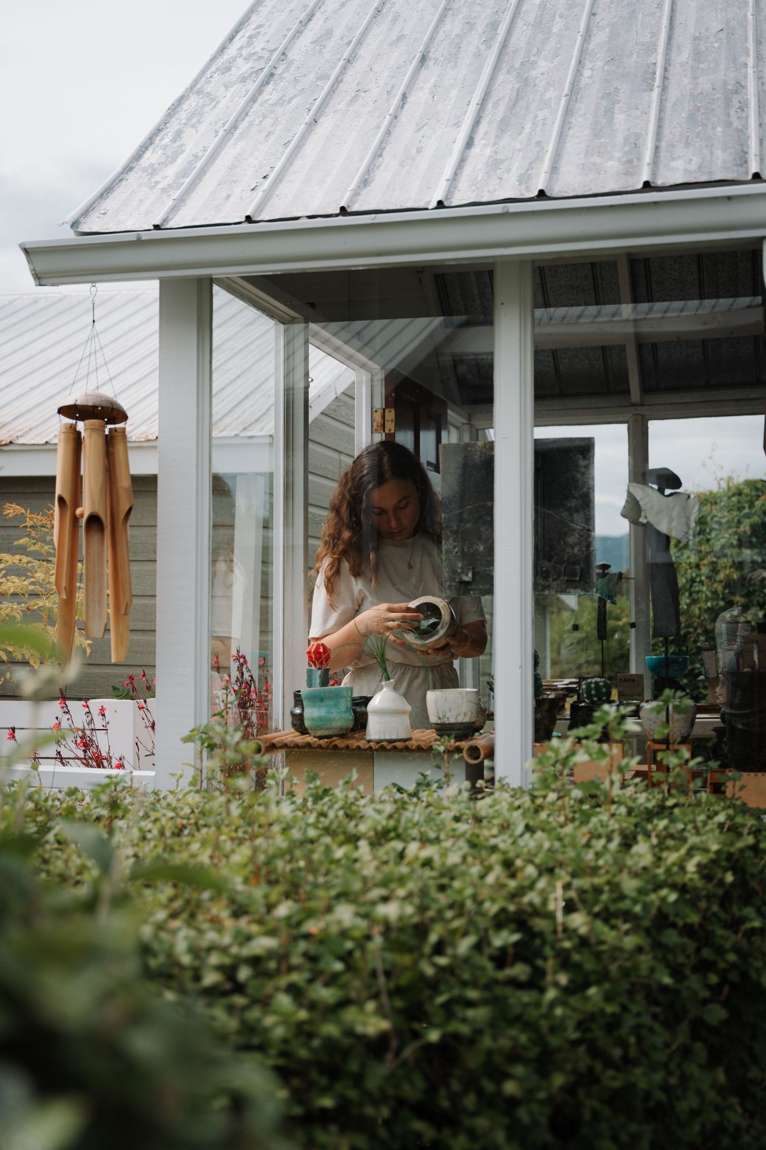 Woman arranging ceramic pieces on a wooden table in a garden greenhouse surrounded by lush green plants.