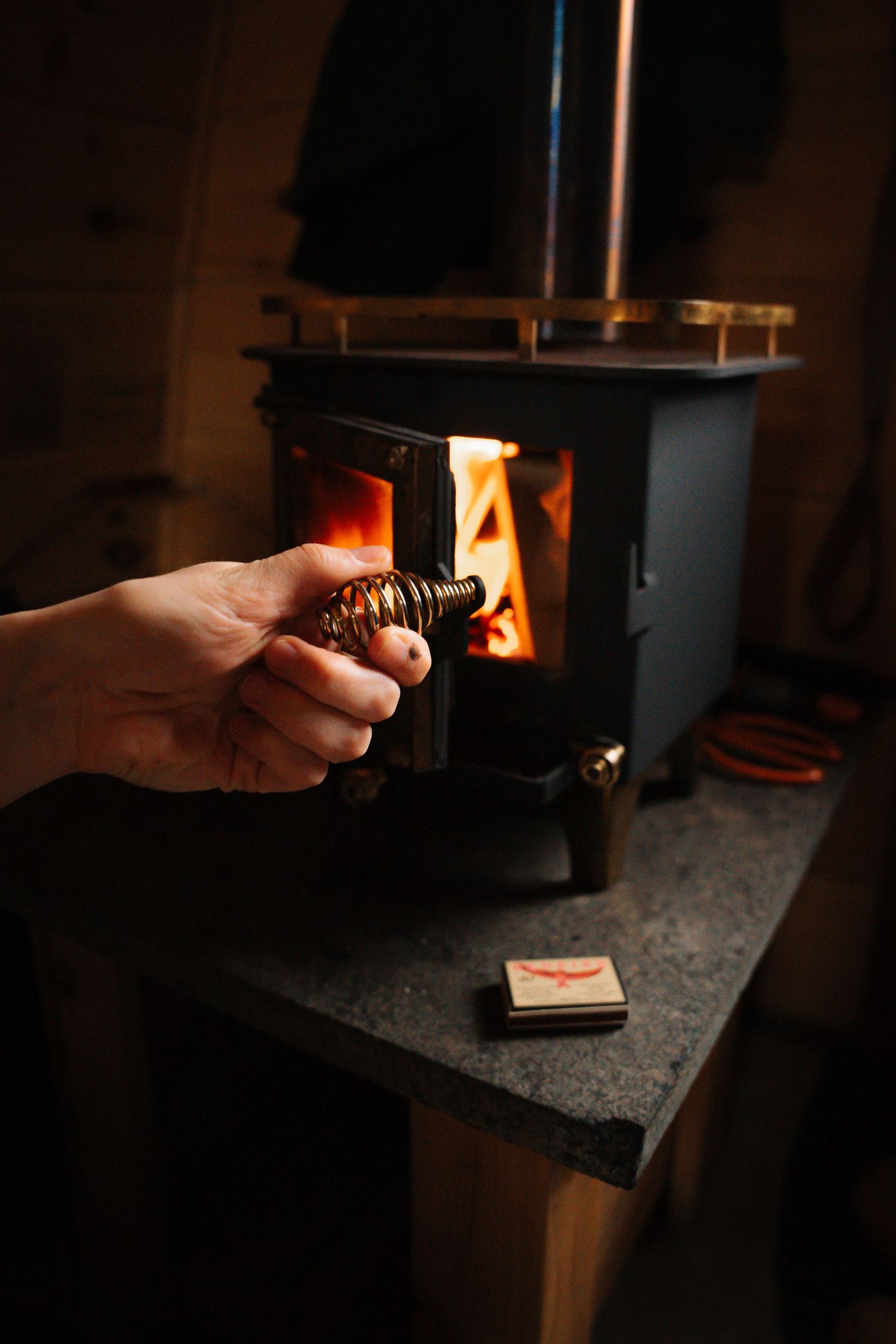 Hand holding a metal coil near an open wood-burning stove with glowing fire inside.
