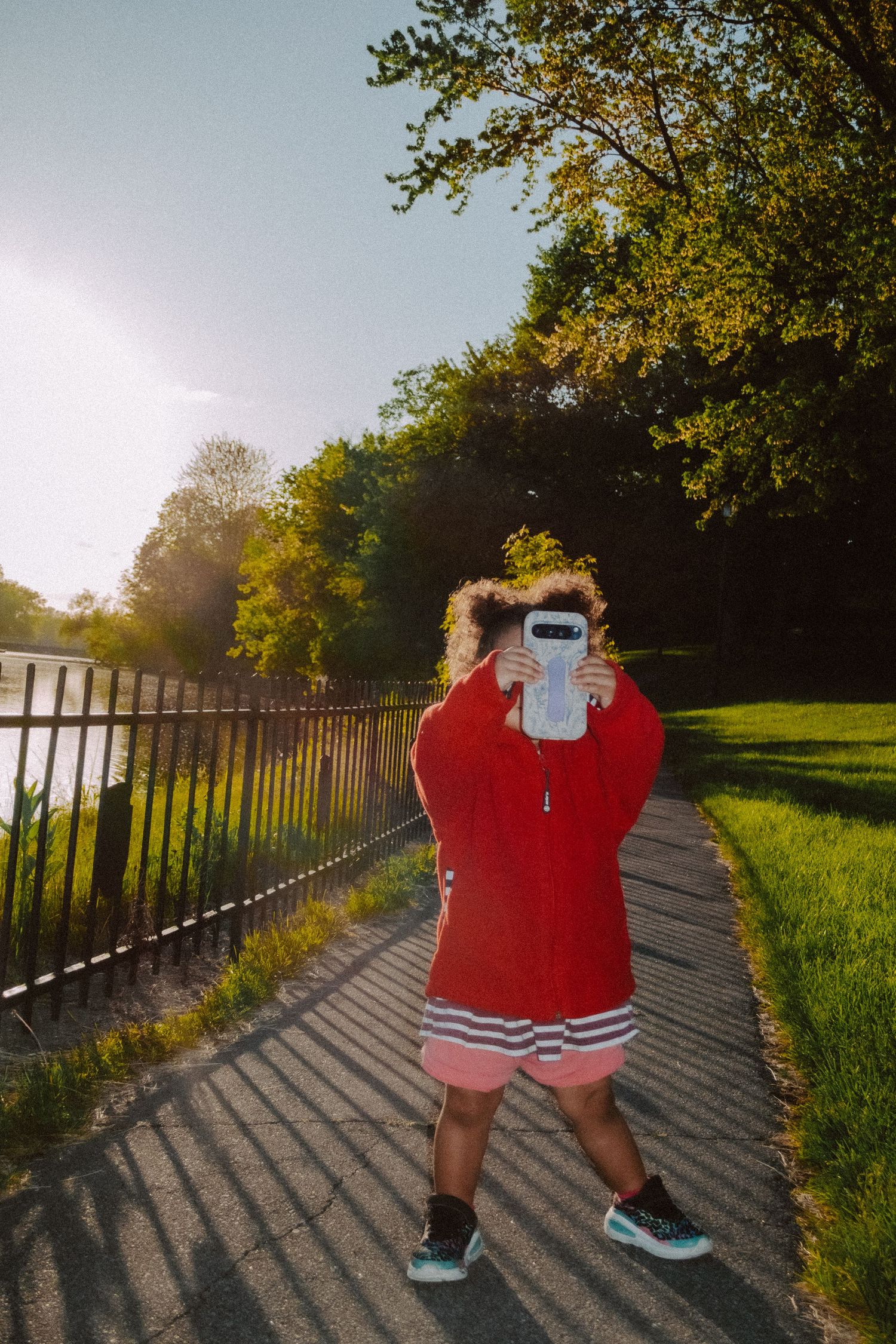 Person in red jacket holding phone over face on sunny park path with green trees and iron fence.