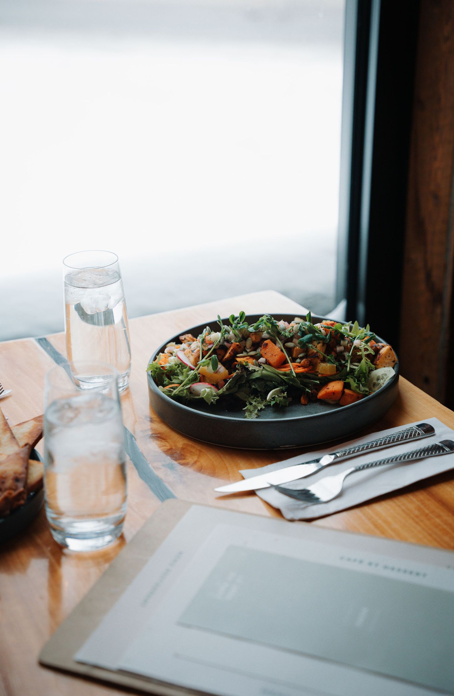 A fresh salad in a dark bowl on a wooden restaurant table with water glasses and silverware by a bright window.