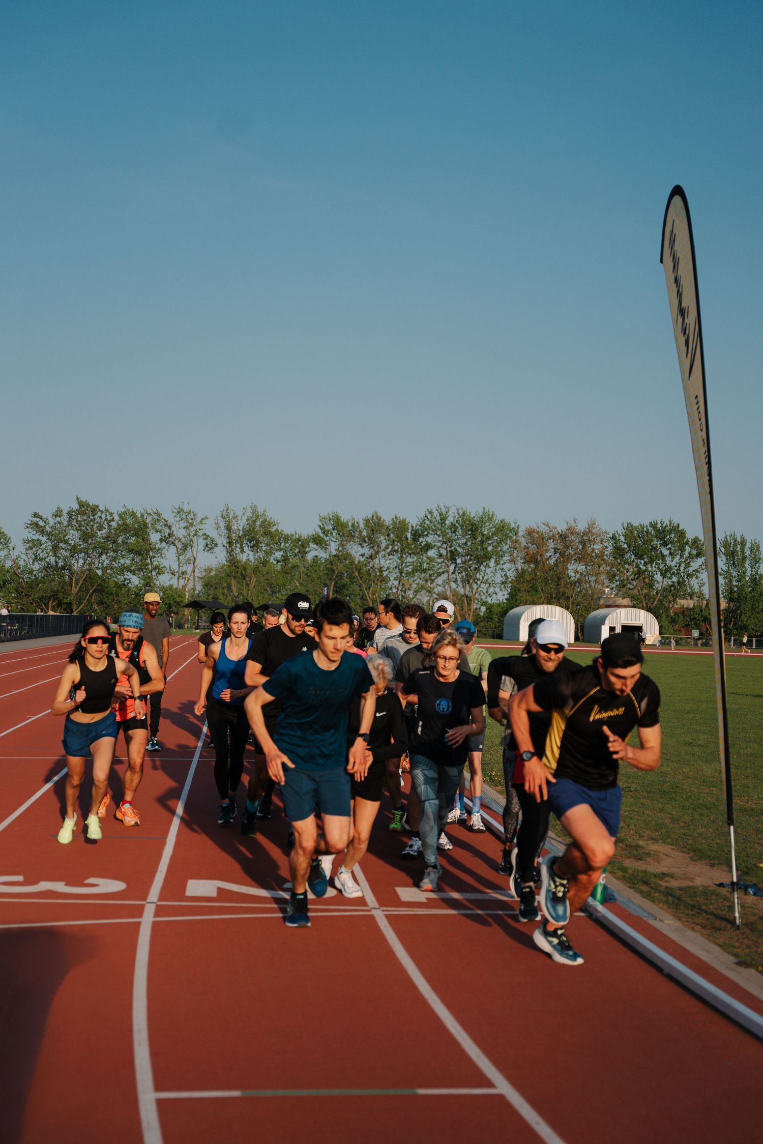A large group of runners races on an outdoor athletic track under a clear blue sky.