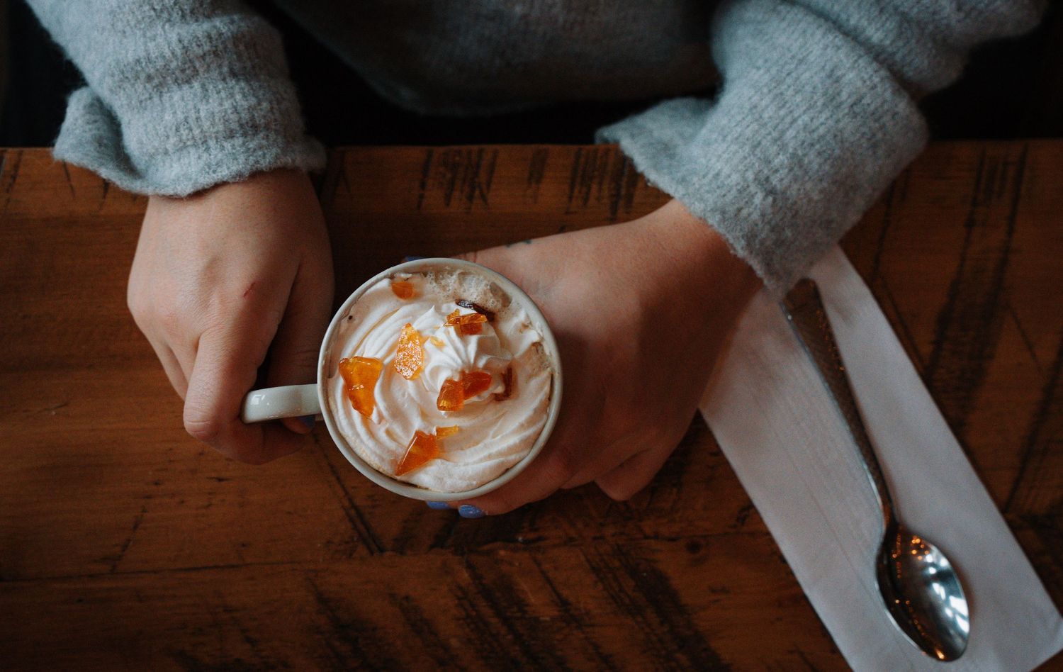 Overhead view of hands holding a white mug of latte with whipped cream and caramel drizzle on wood table.