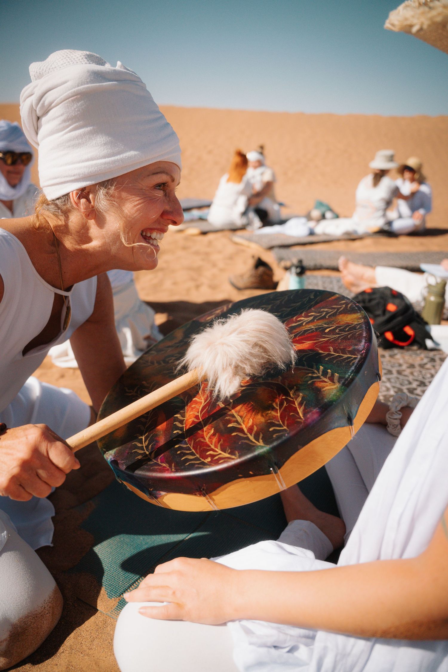 Smiling woman in white turban playing a decorative shamanic drum in the desert.