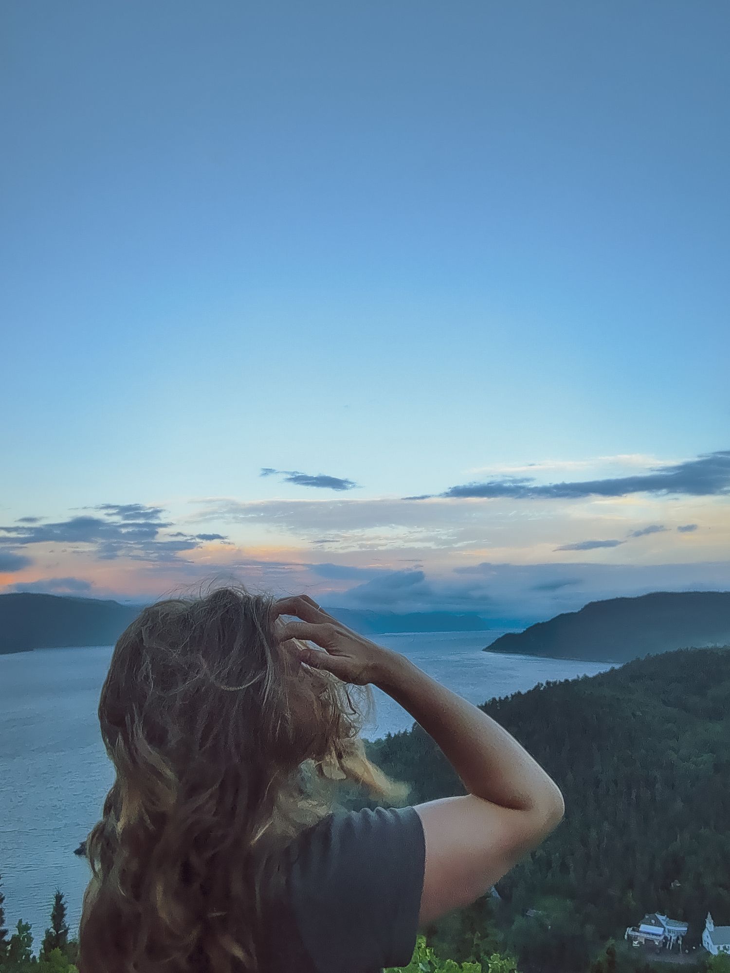 A woman with long wavy hair gazes at a stunning sunset over a fjord from a scenic hilltop viewpoint.