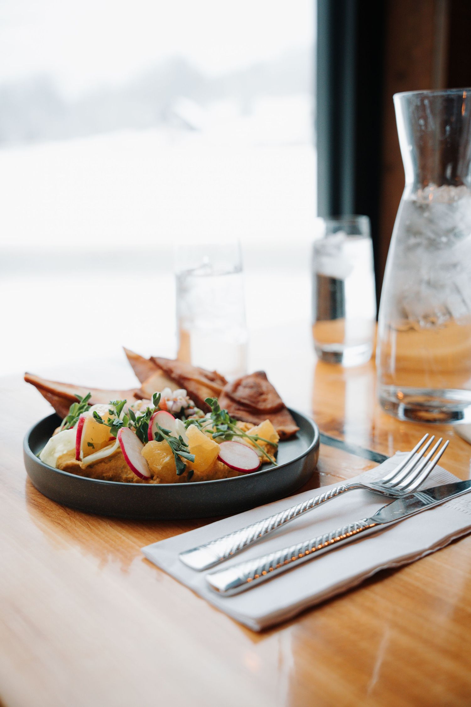 Dark plate with fresh salad, radishes, and crispy flatbread served beside silverware and water carafe.