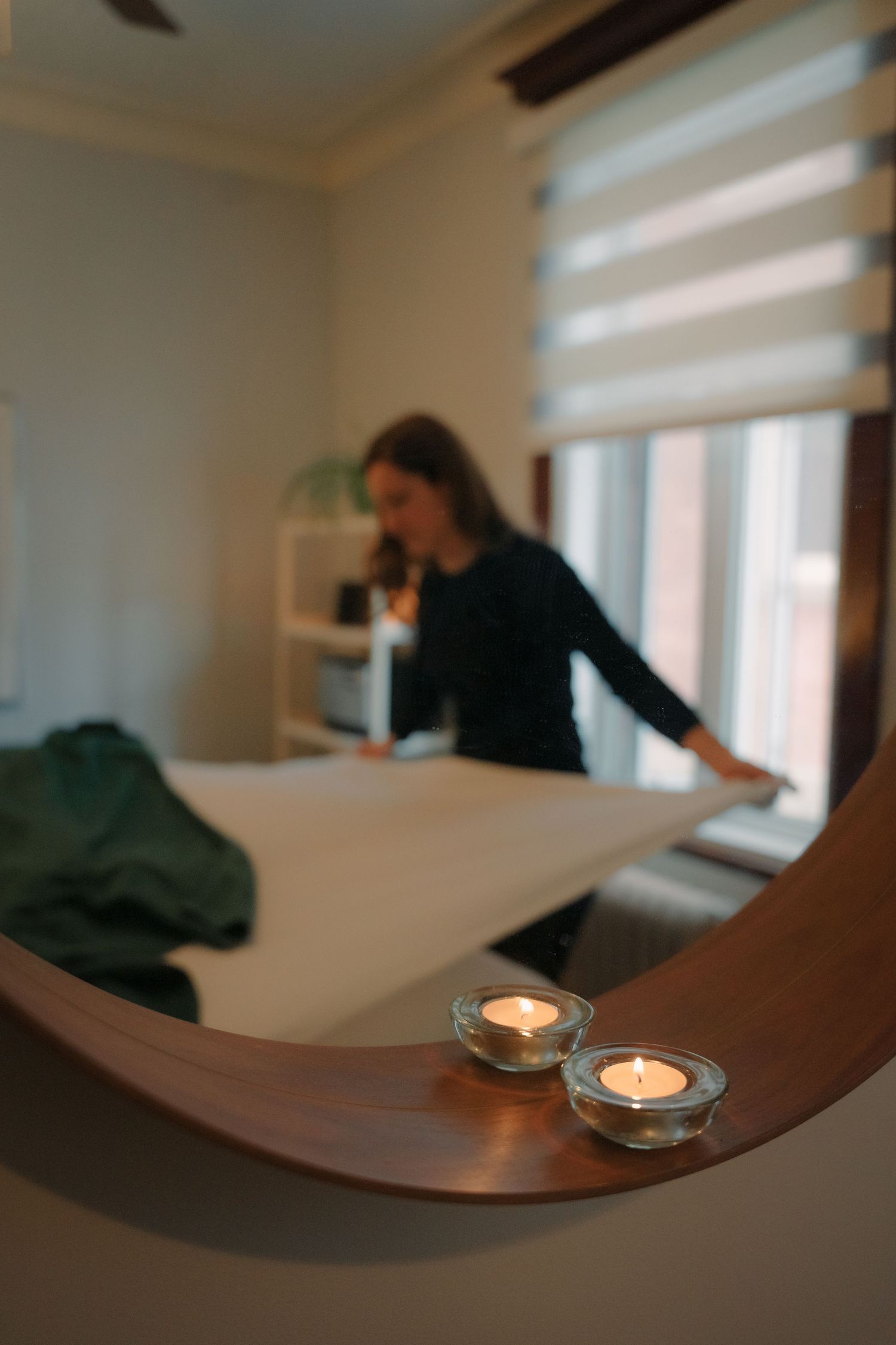 Massage therapist setting up white treatment table with candles in foreground of cozy spa room.