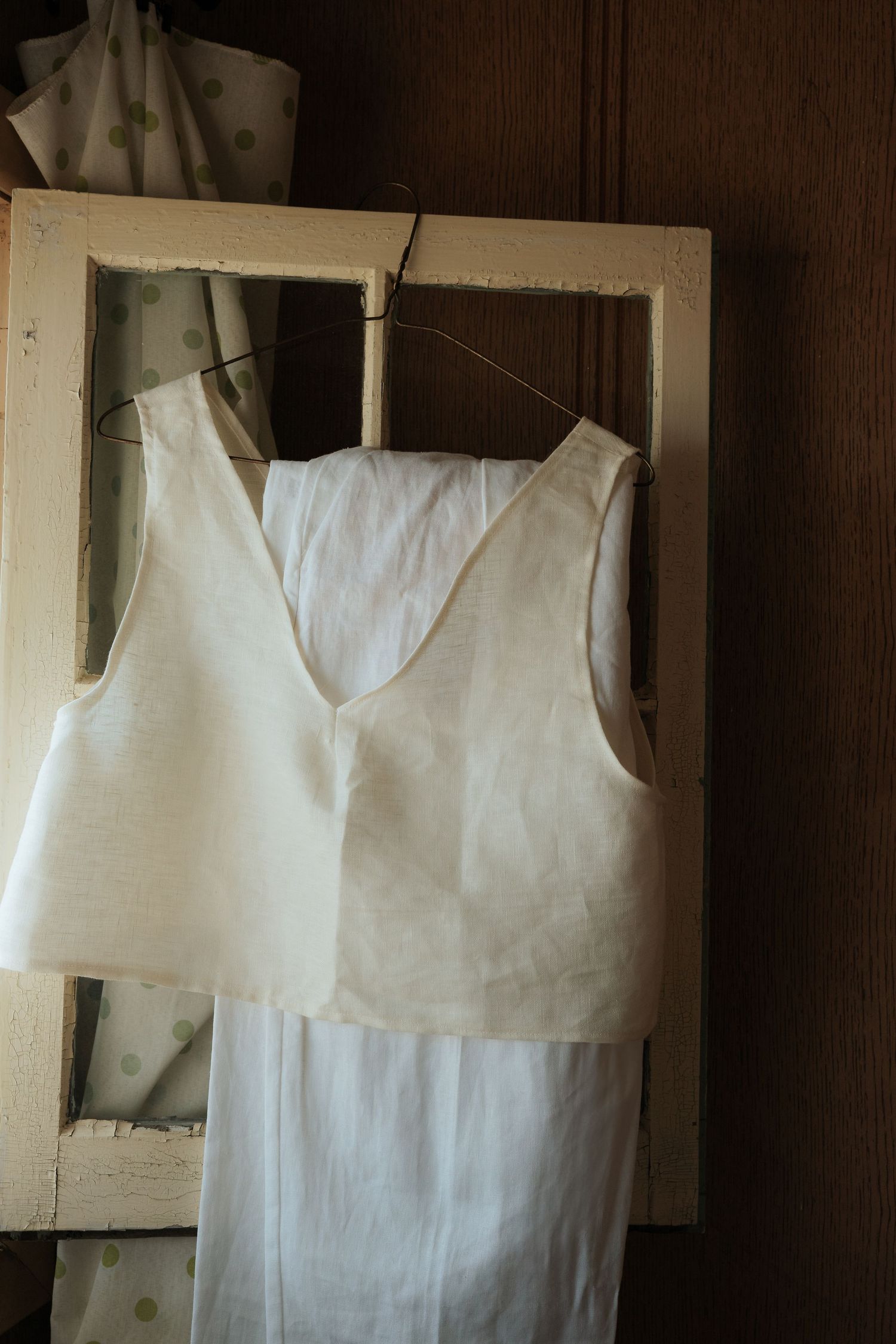 A white sleeveless wedding dress hangs on a vintage frame hanger against a dark wooden wall.