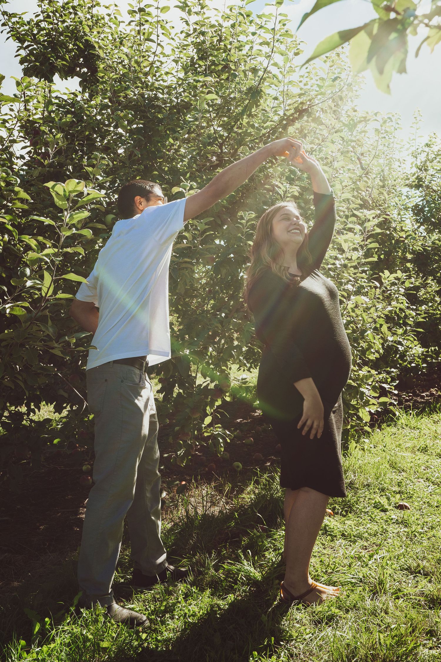 A couple dancing joyfully in a sunlit green orchard, woman visibly pregnant.