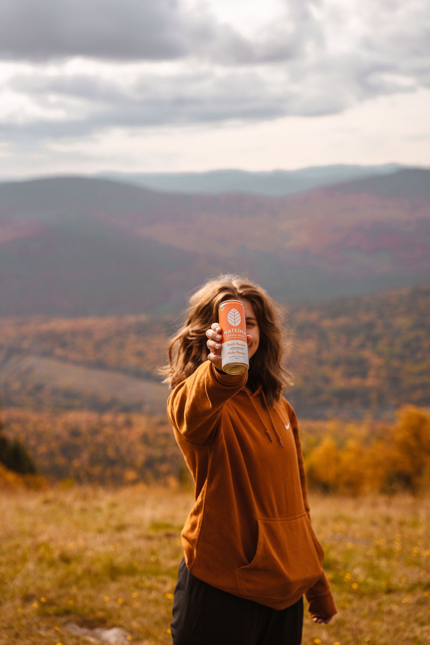 Woman in orange hoodie holding a can toward camera with autumn mountain landscape behind her.