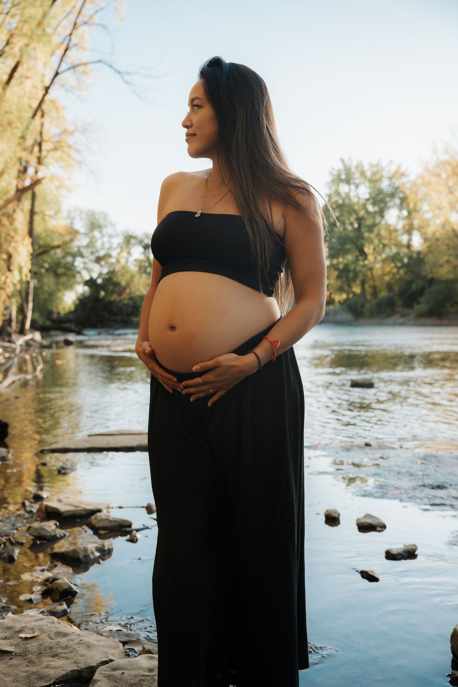 A pregnant woman in black holds her belly while standing beside a peaceful rocky river.