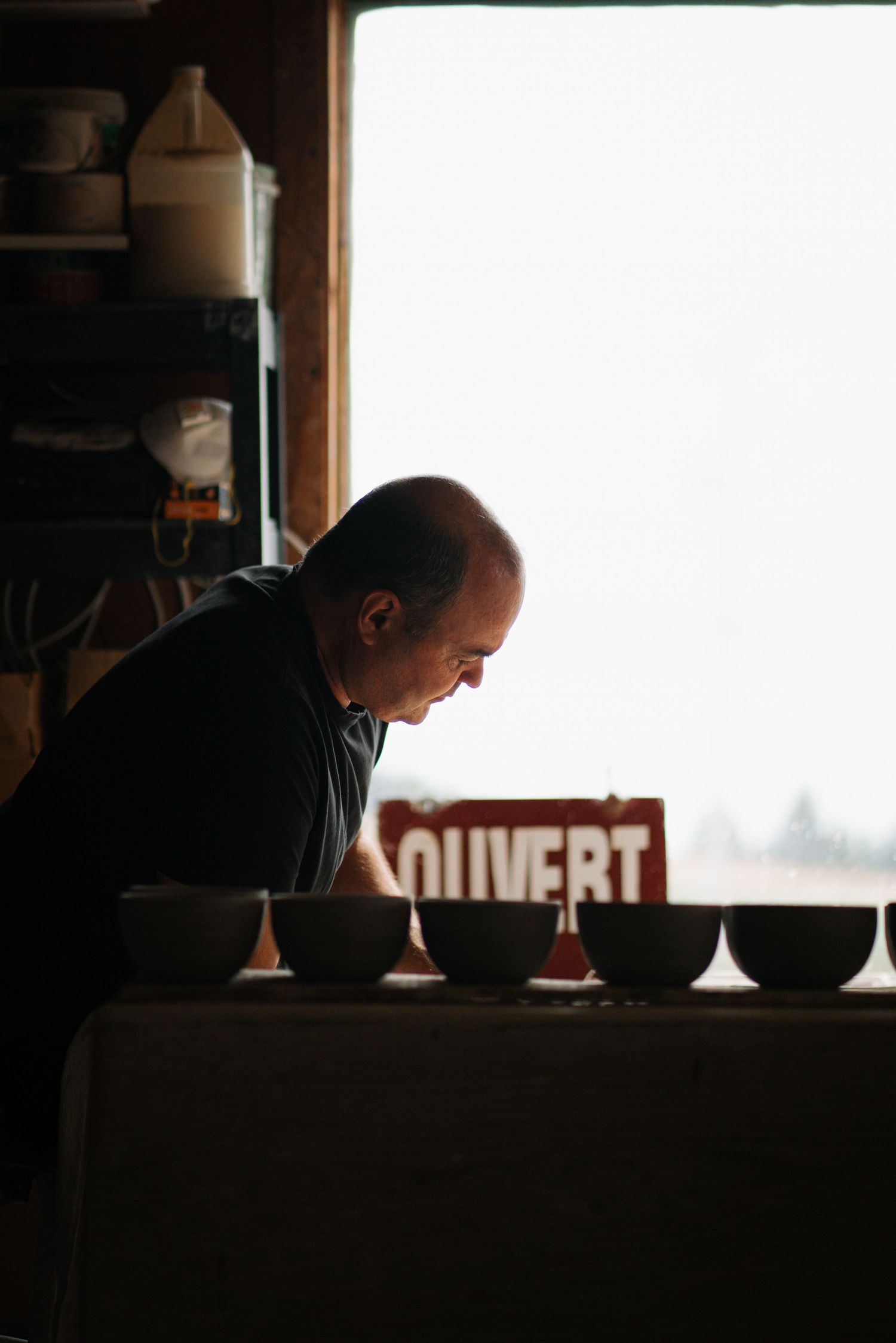 A bald man leans over a table of ceramic bowls in a dimly lit pottery studio with an OUVERT sign.