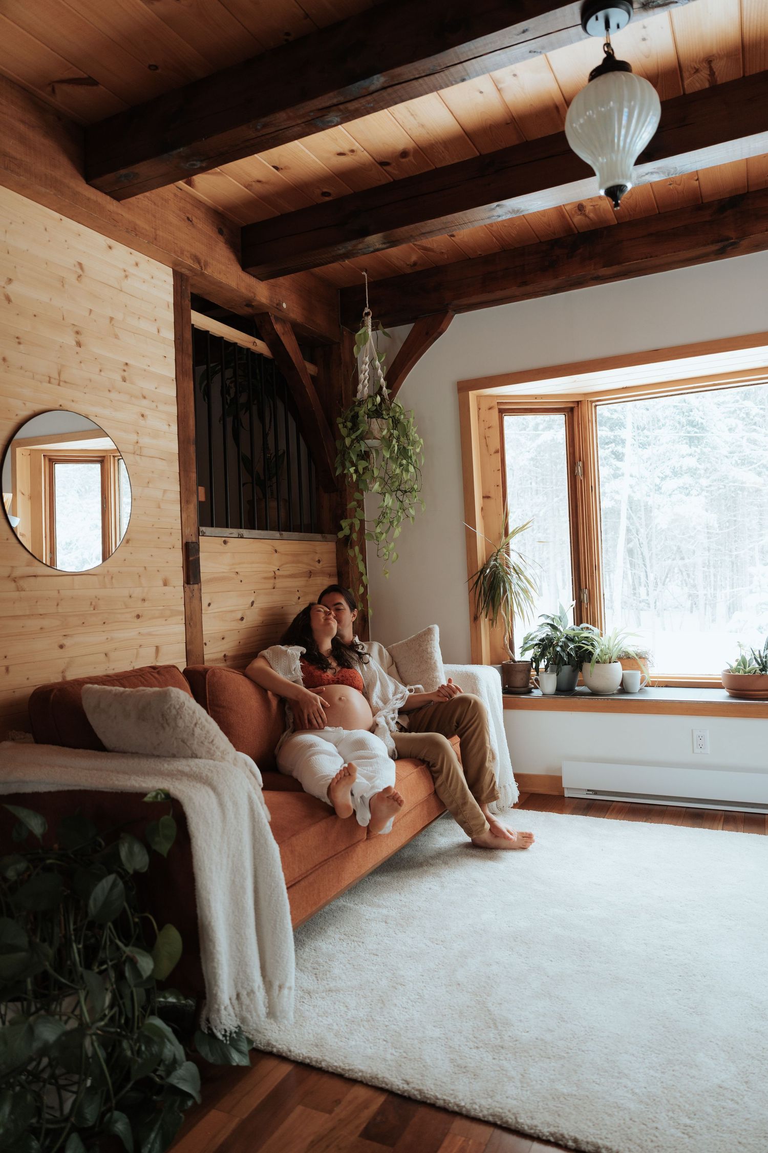 Woman relaxing on orange sofa in cozy wooden cabin living room with plants and snowy window view.