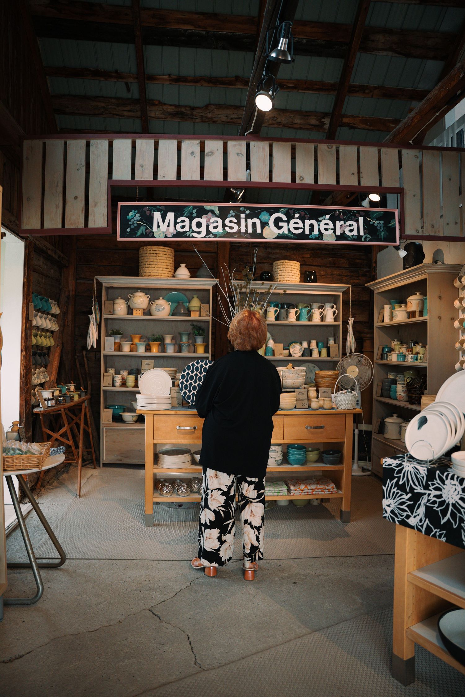 Customer browsing handmade ceramics at a rustic Magasin General shop with wooden shelving displays.